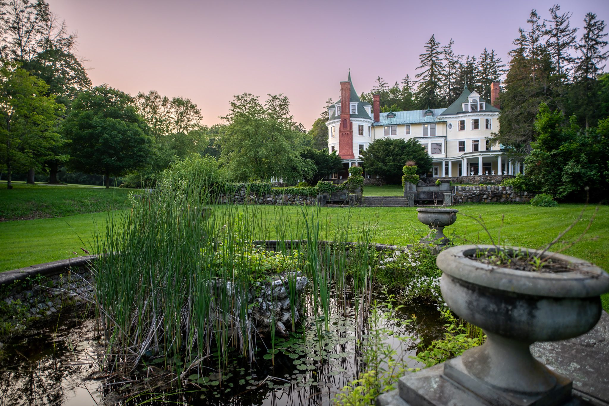 From the edge of the fountain pool, reeds and a stone urn in the foreground and the house under a lilac sky.