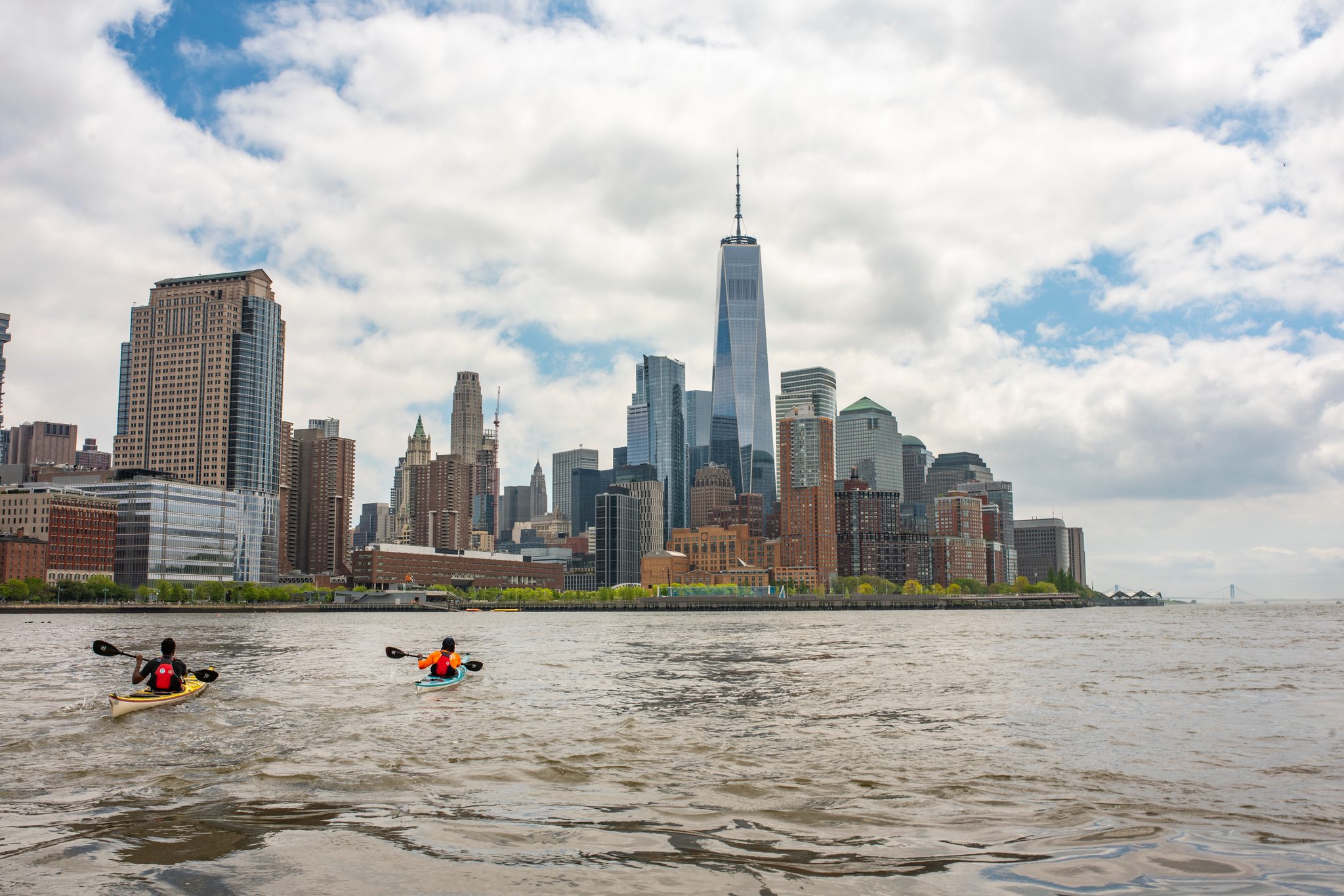Two tiny boats on open water at the tip of Manhattan, the skyline heavy and bright above them.