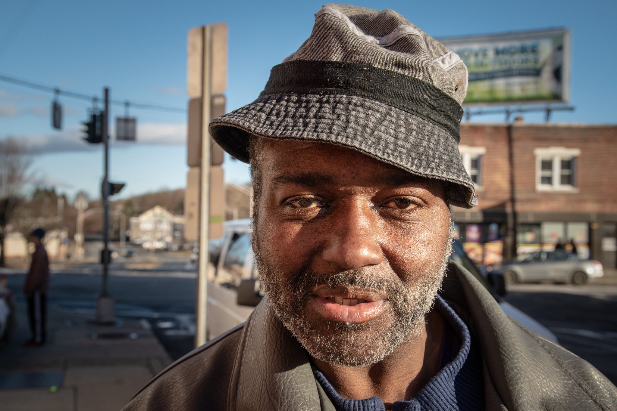 A man in a worn bucket hat pauses on the corner long enough to be seen.