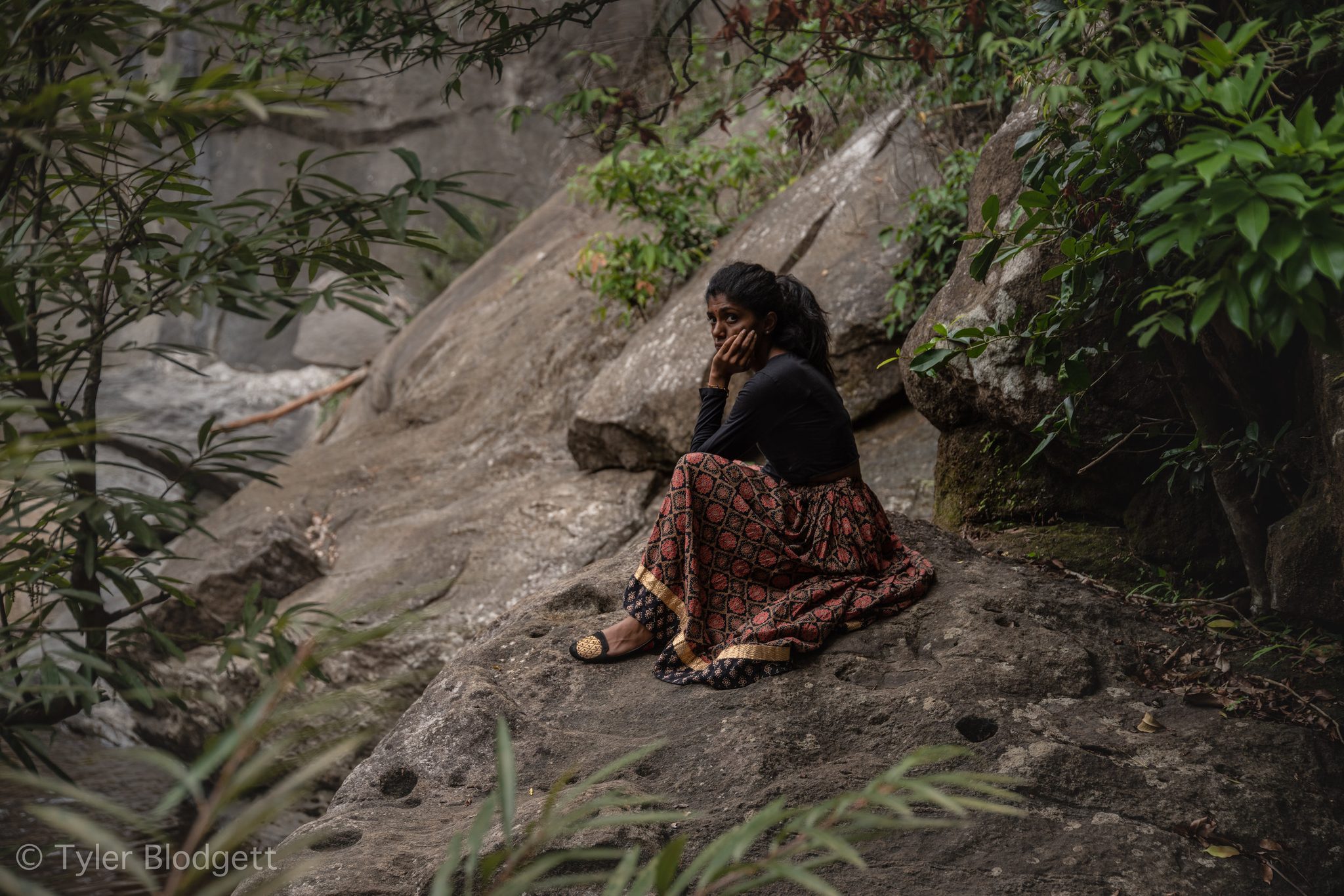 A young woman sits on the rock face, chin in hand, the forest all around her.