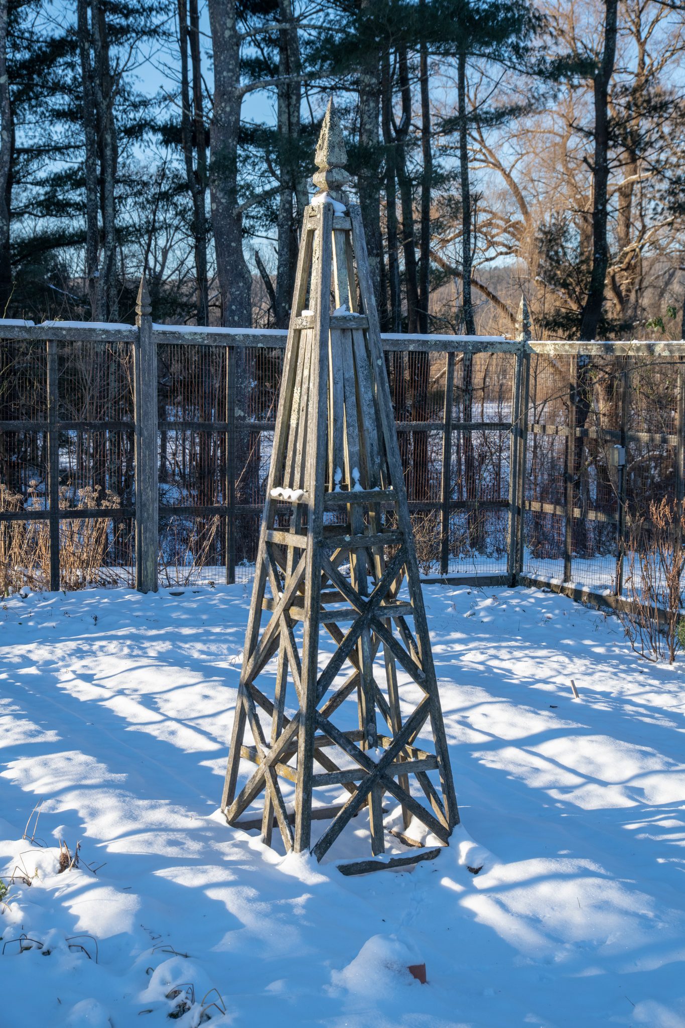 A garden obelisk stands alone in the snow, the fence and pines behind it.