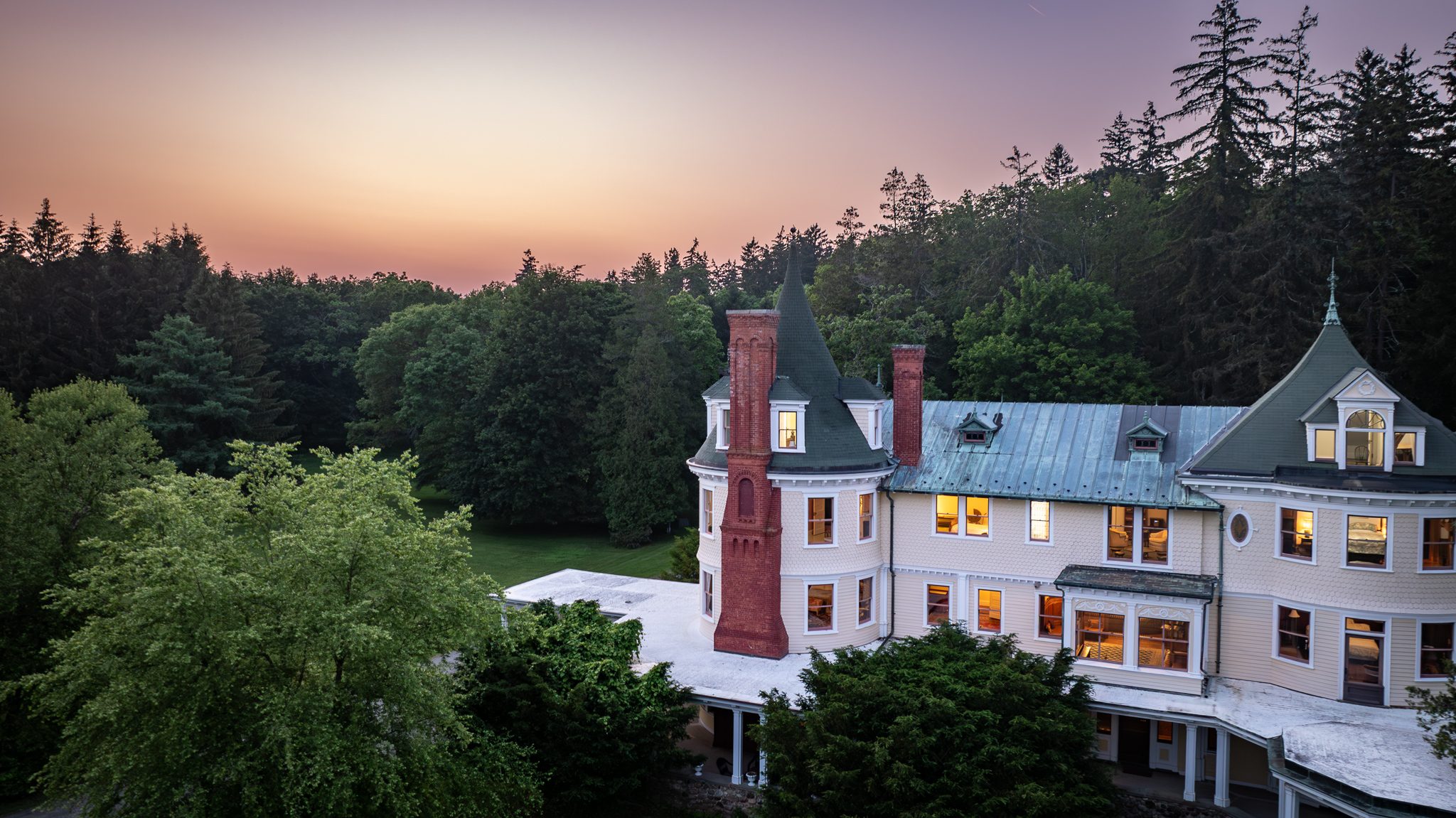 Lamps coming on inside as the sky over the pines turns orange, the brick chimney still holding a little color.