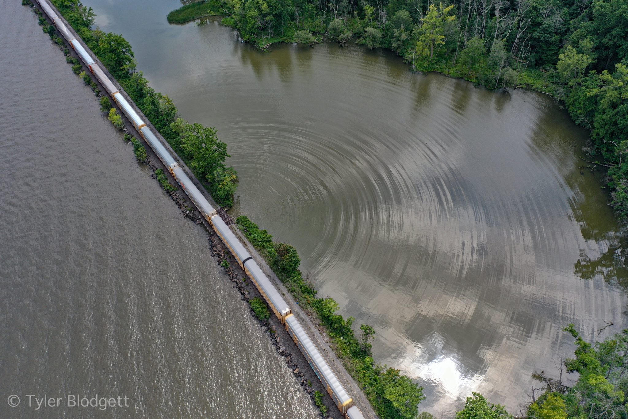 A freight train threads the causeway between the river and a still cove.