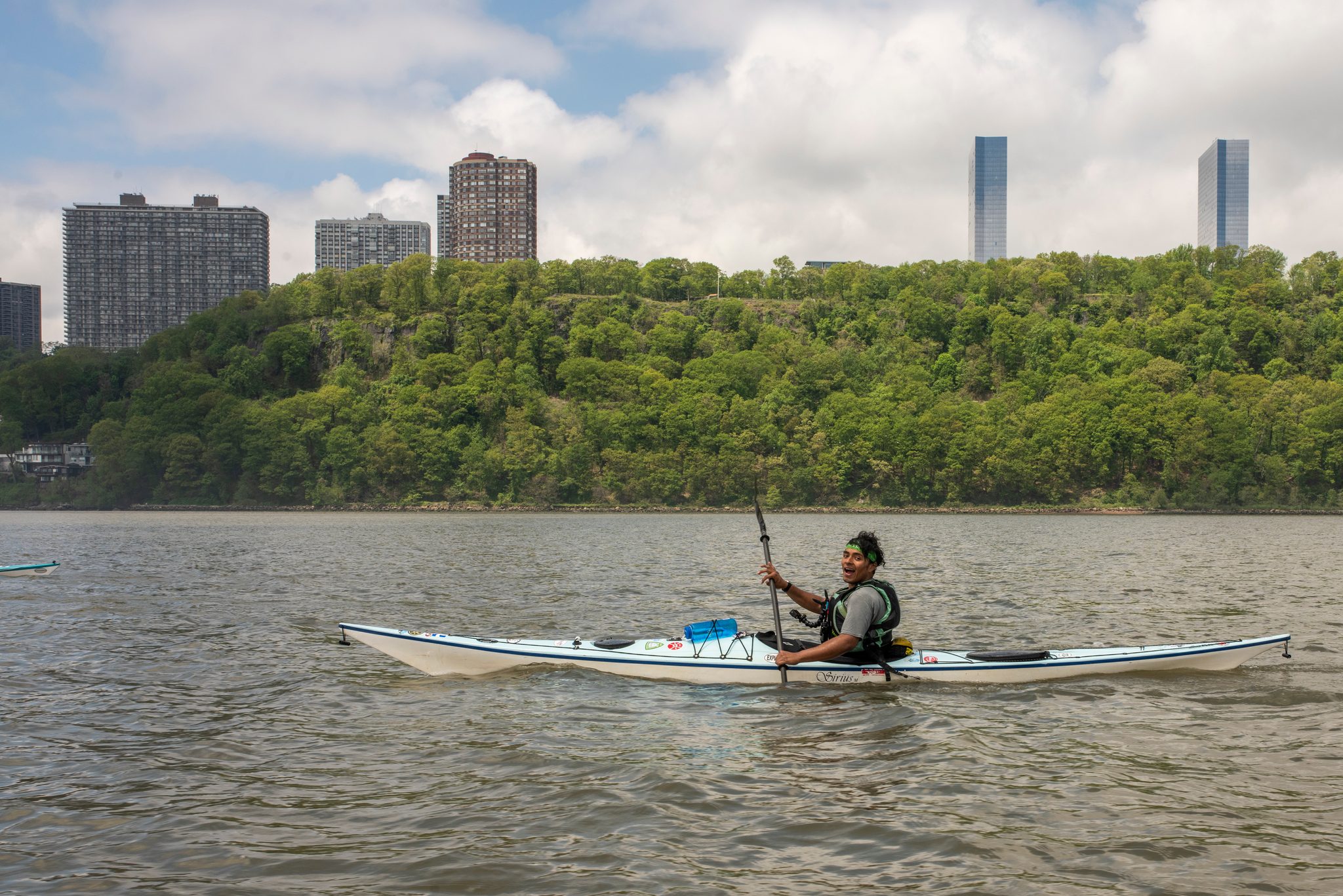 A single kayak skims past a green bluff crowned with apartment towers on the Manhattan side.