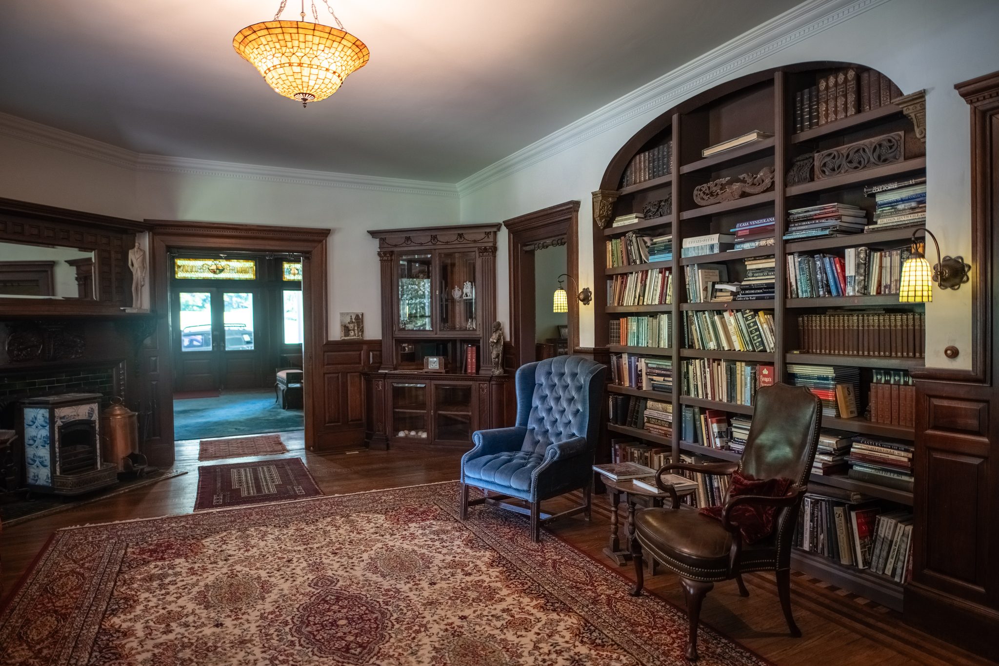 A library of arched built-in shelves, a blue wingback by the window and a leaded-glass shade glowing overhead.