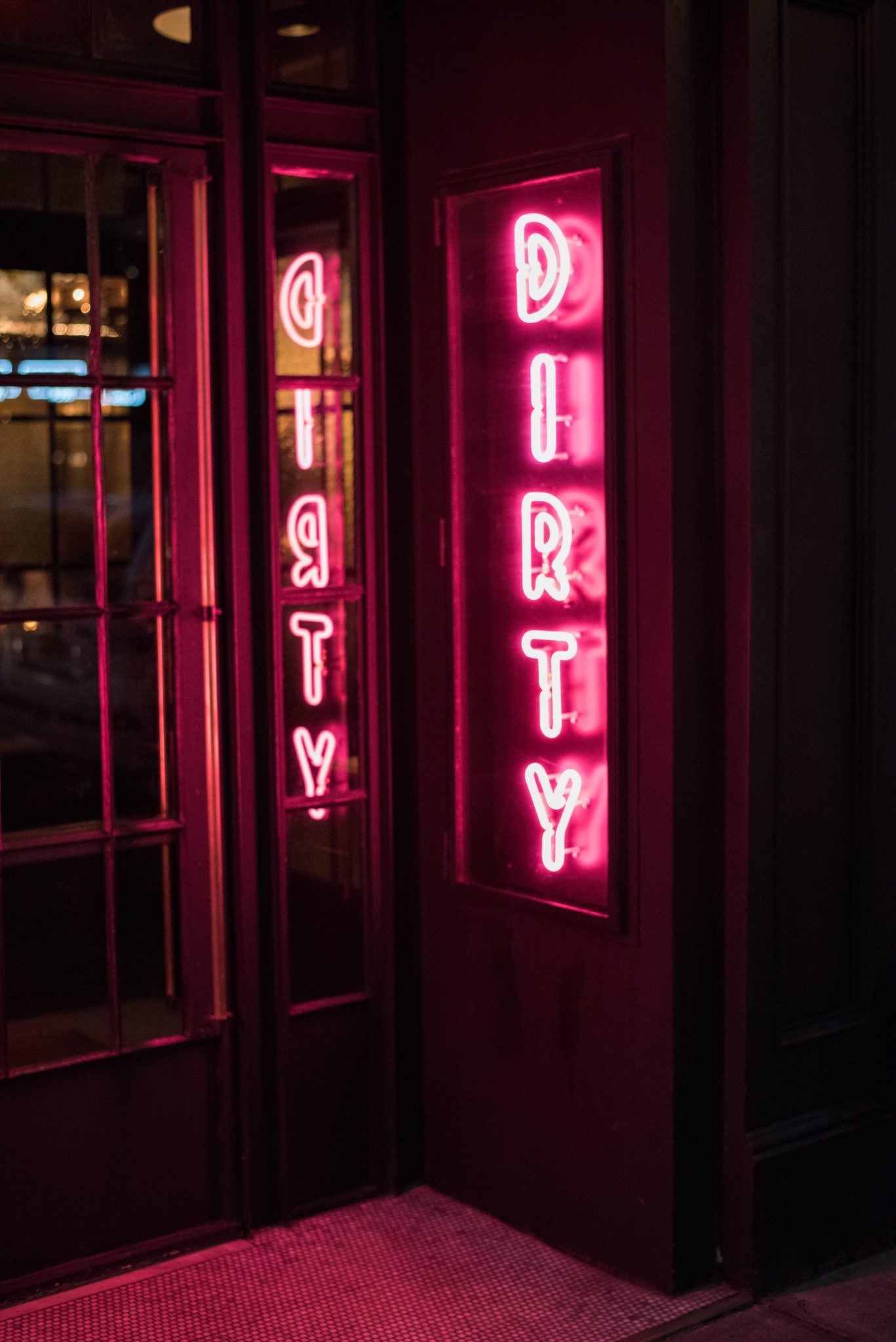 The word DIRTY burns pink in neon, doubled in the glass of the door.