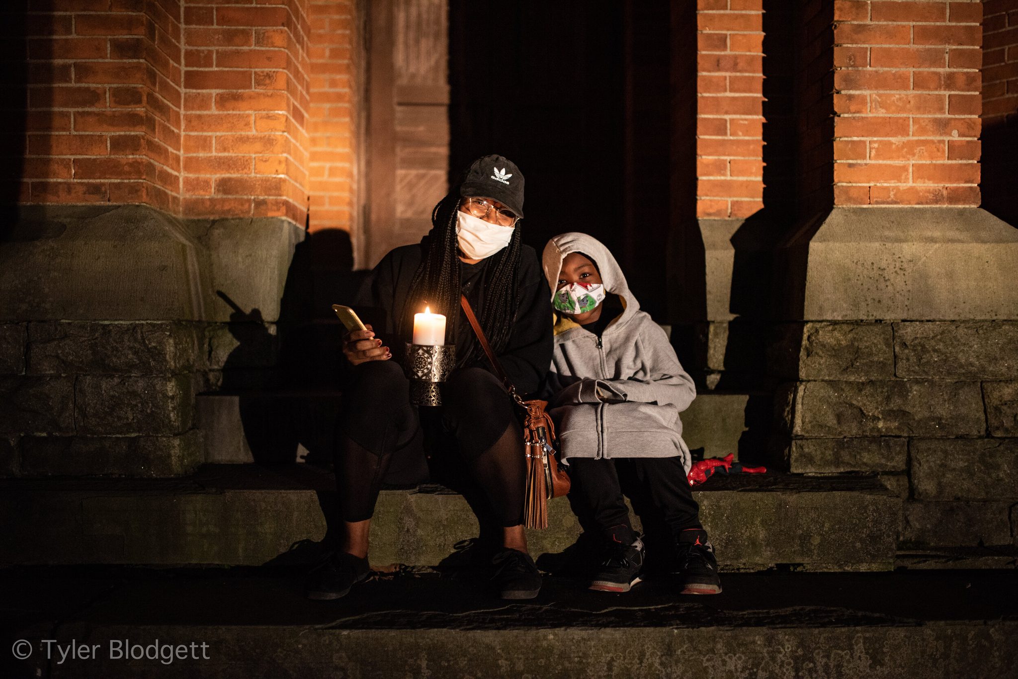 Two mourners sit on the church steps, candles in hand, masks on.