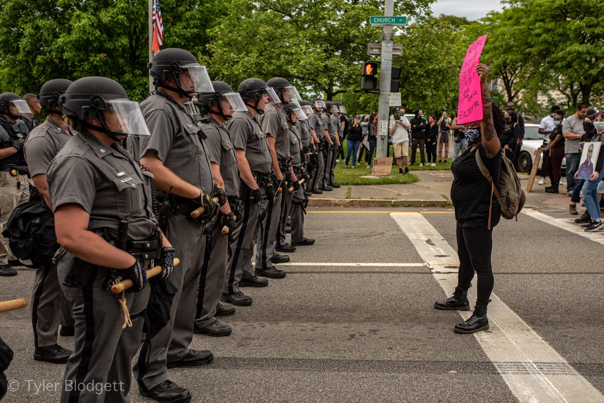 A woman stands alone in the crosswalk facing a line of troopers, her pink sign held high.