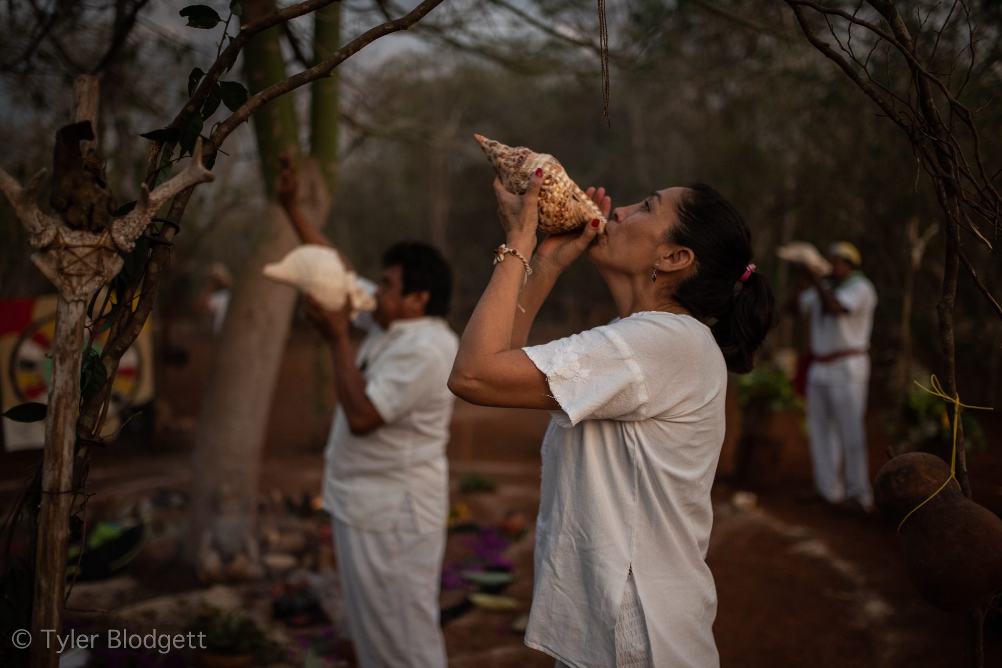 A woman in white blows the conch, head tipped back.