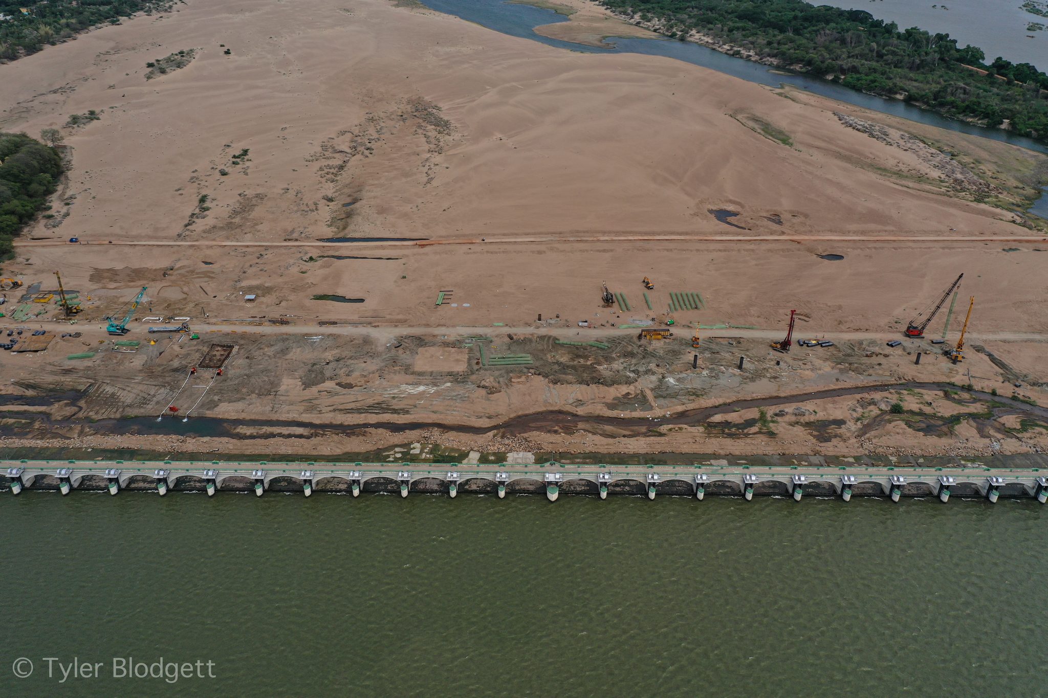 Construction crews work the sand beside a long bridge over the river.