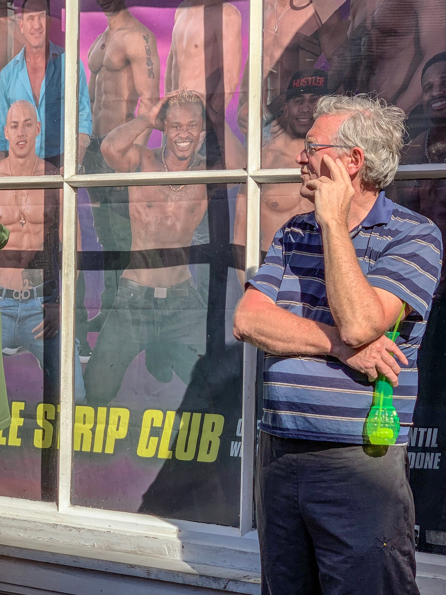 A man in a striped polo stops to think, drink in hand, in front of a strip club window.