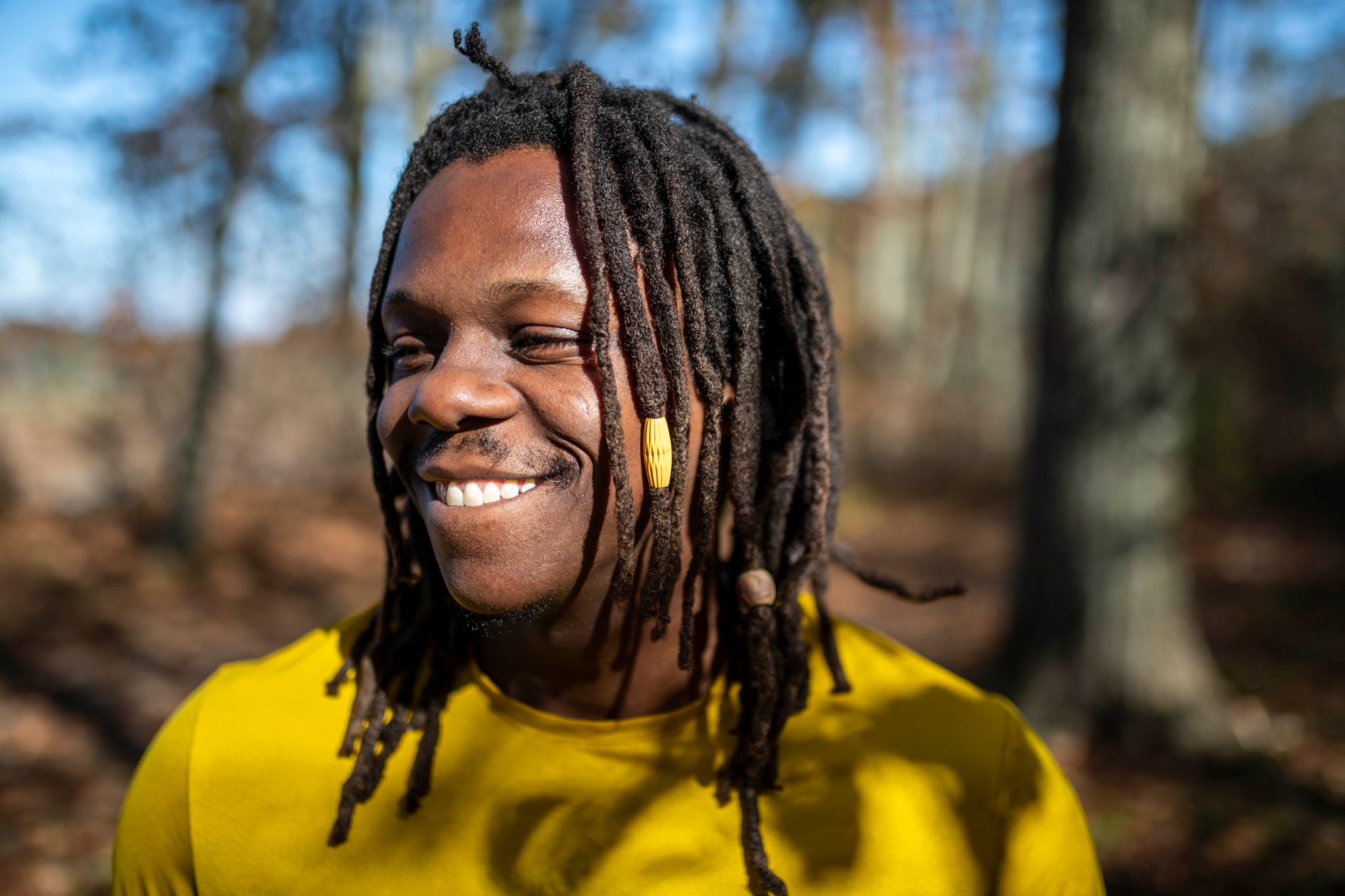 Chev laughs in a yellow shirt among autumn trees, sun catching the beads in his locs.