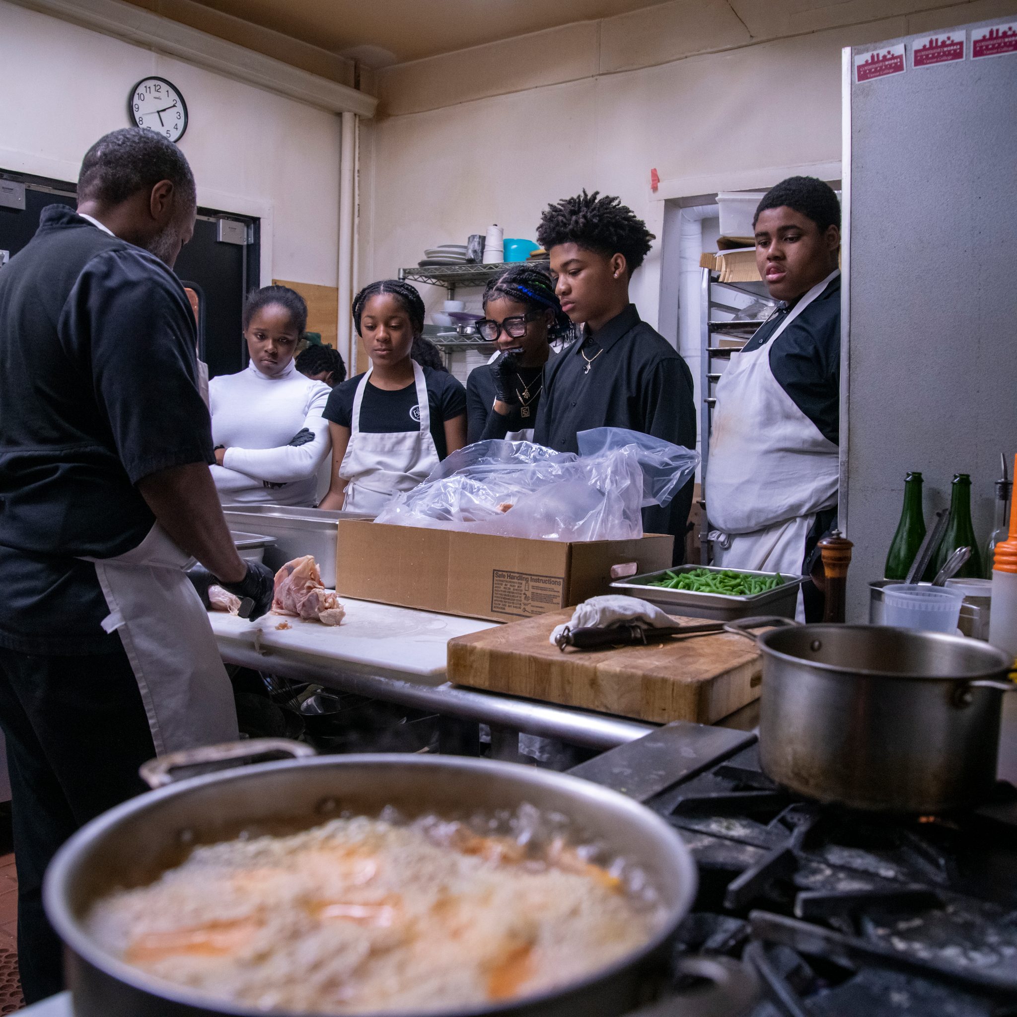 A pot bubbles in the foreground while the chef shows the young cooks the next step.