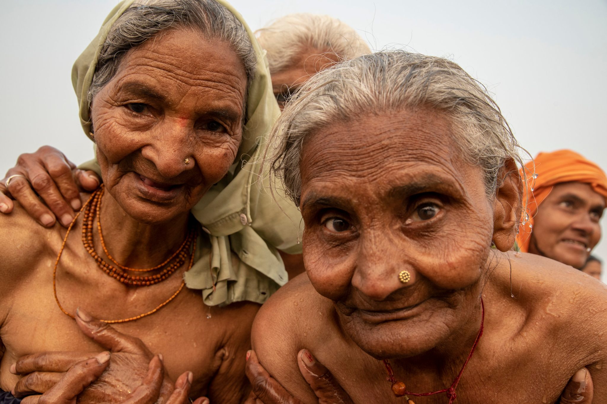 At the Kumbh Mela, two elder women press close to the camera, hair still wet from the river, their hands on each other's shoulders.