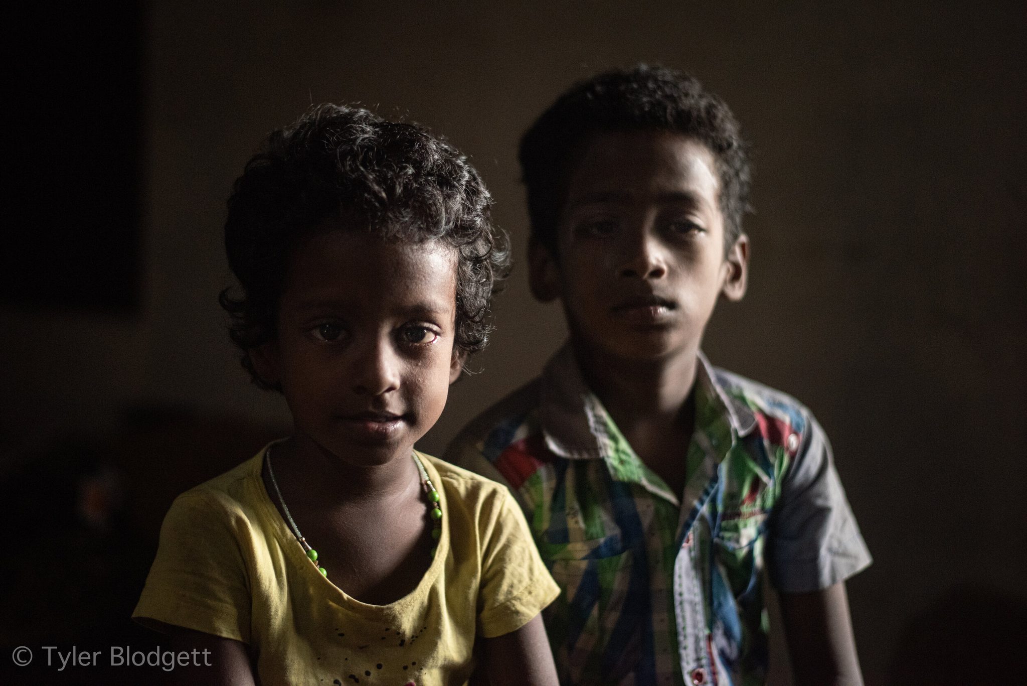 Two boys hold still in the low light, the younger one meeting the lens straight on.