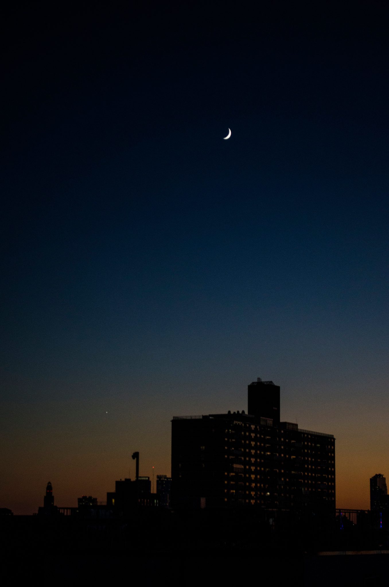 A crescent moon hangs over the rooftops as the last orange light fades.