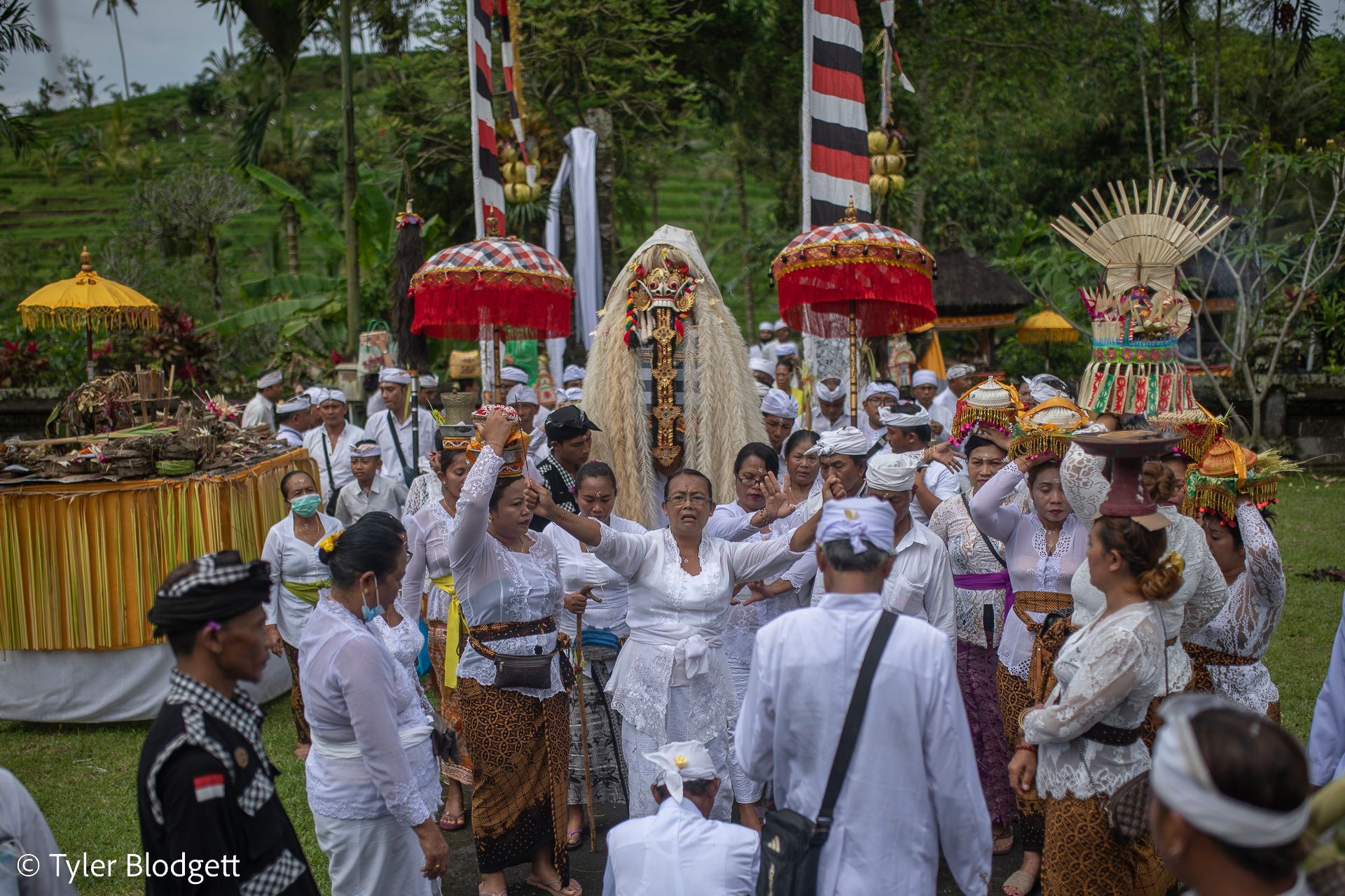 The procession moves out beneath fringed parasols, offerings balanced on heads and a masked figure towering over the crowd.