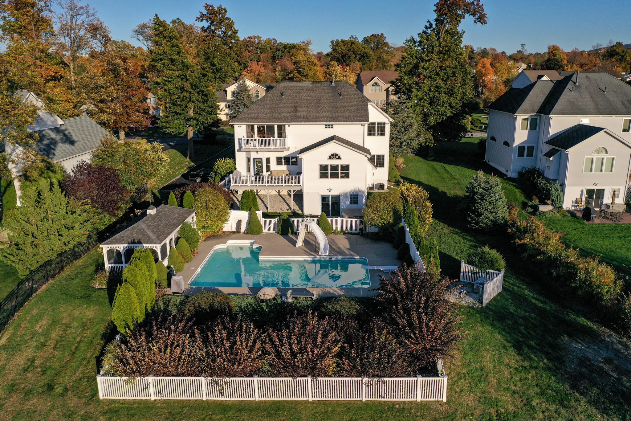 A backyard pool sits behind a white picket fence, the neighborhood in full autumn around it.