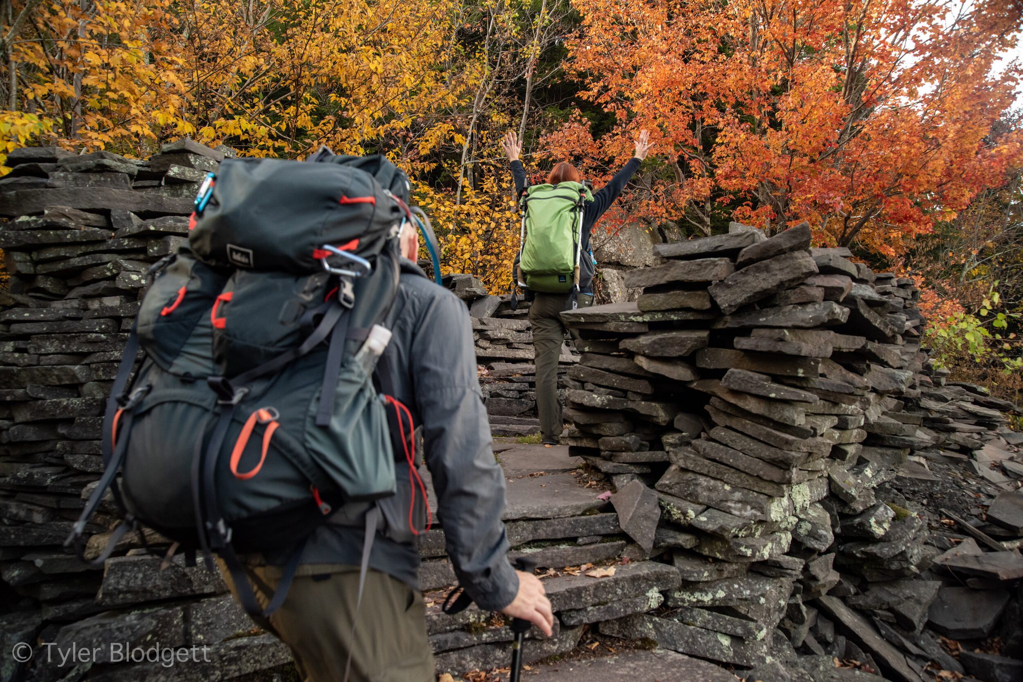 Backpacks climb through stacked slate and blazing autumn leaves, and the one out front throws his arms up at the top.