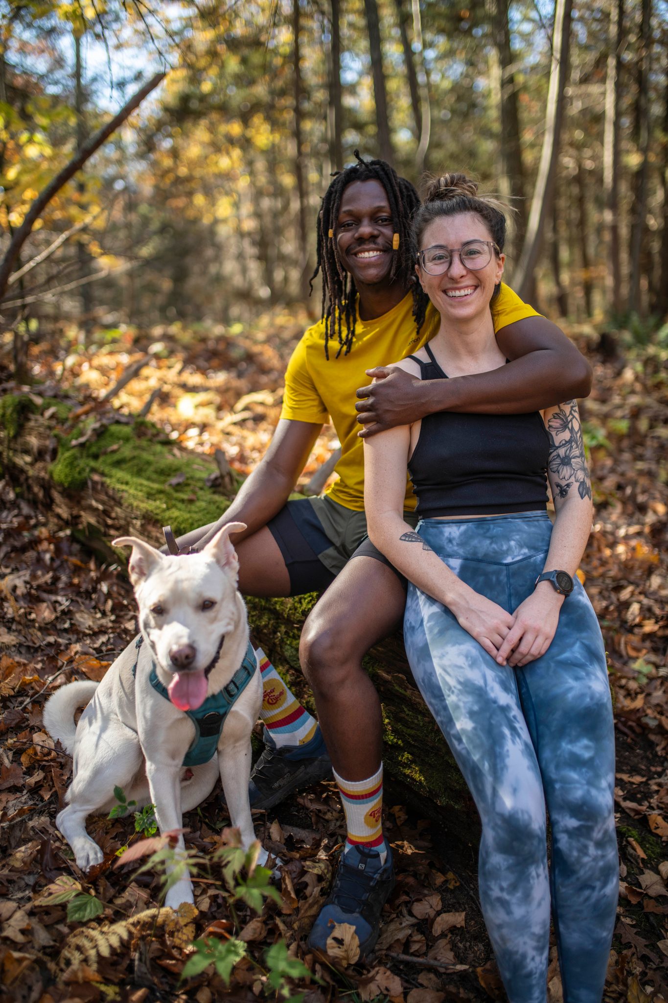 Two friends sit on a mossy log with their white dog, arms around each other.