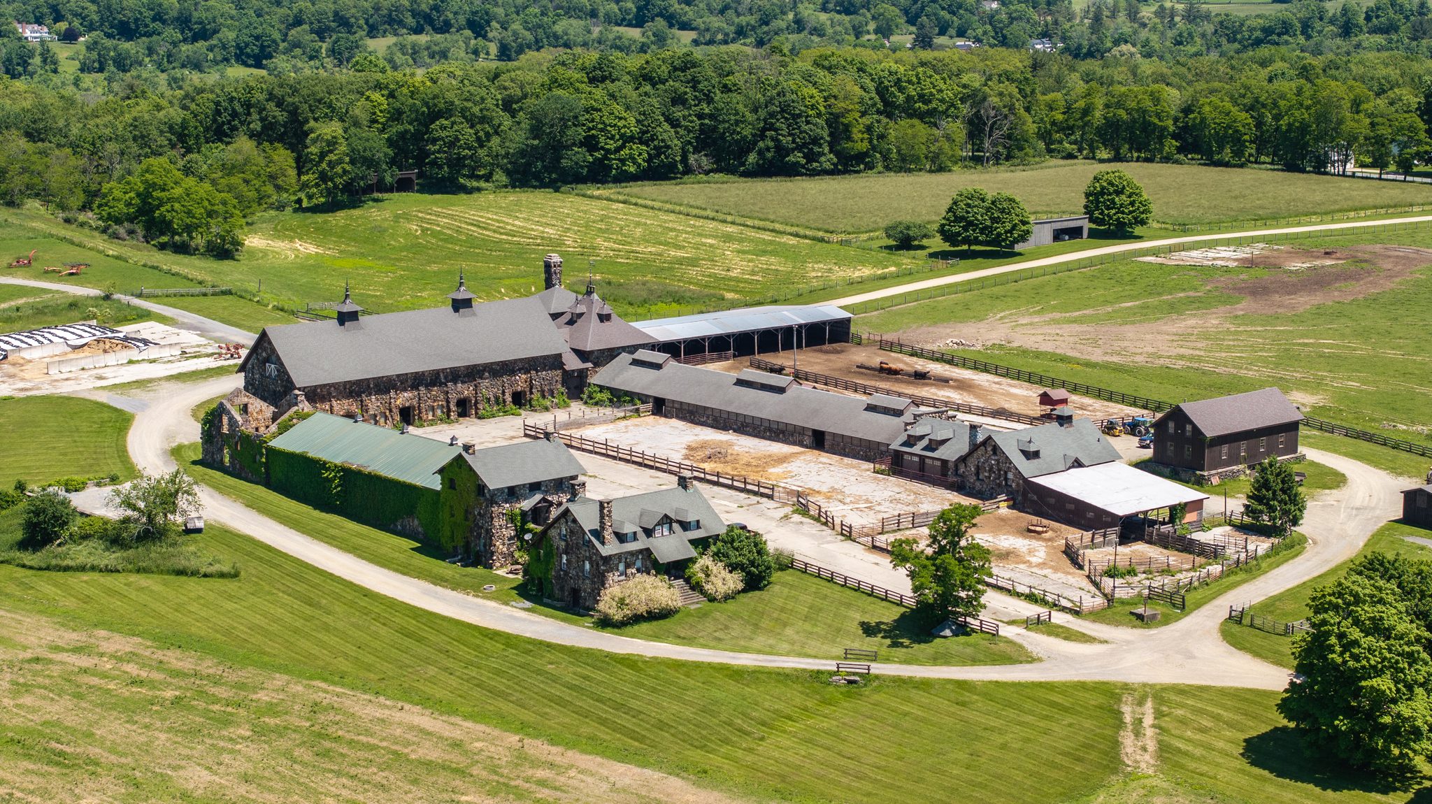 The stone barn complex from the air, long slate roofs and cupolas ringed by paddocks, fencing and mown fields.
