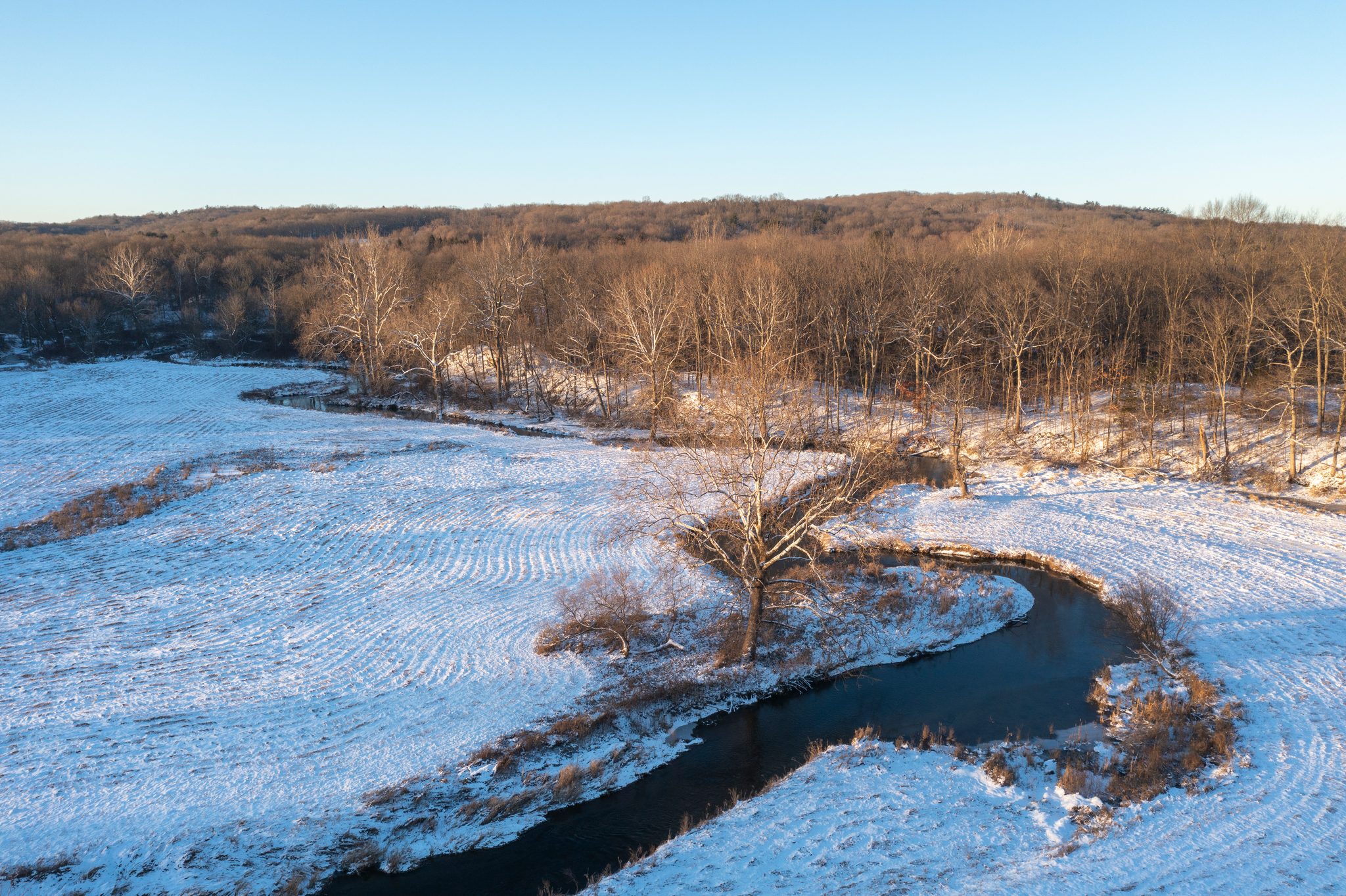Snow on the lower meadow, the stream cutting a black curve through it.