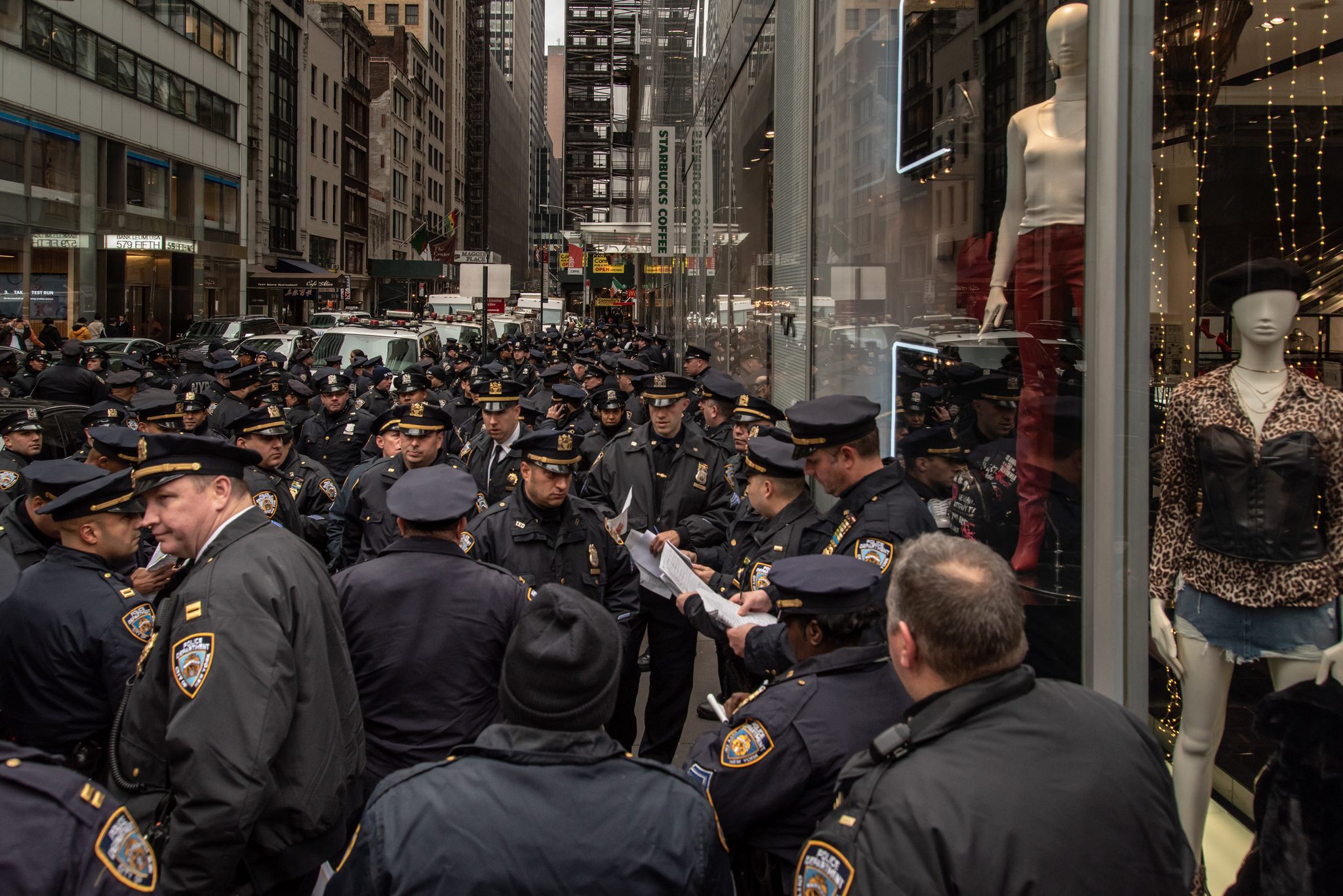 A sea of NYPD caps fills the sidewalk while mannequins look on from the shop window.