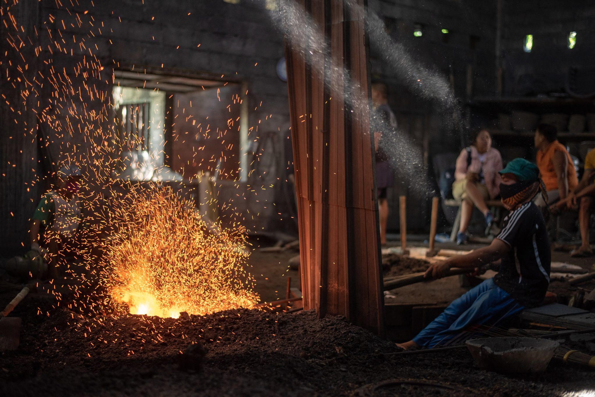 A masked worker tends the forge as sparks leap into the dark of the workshop.
