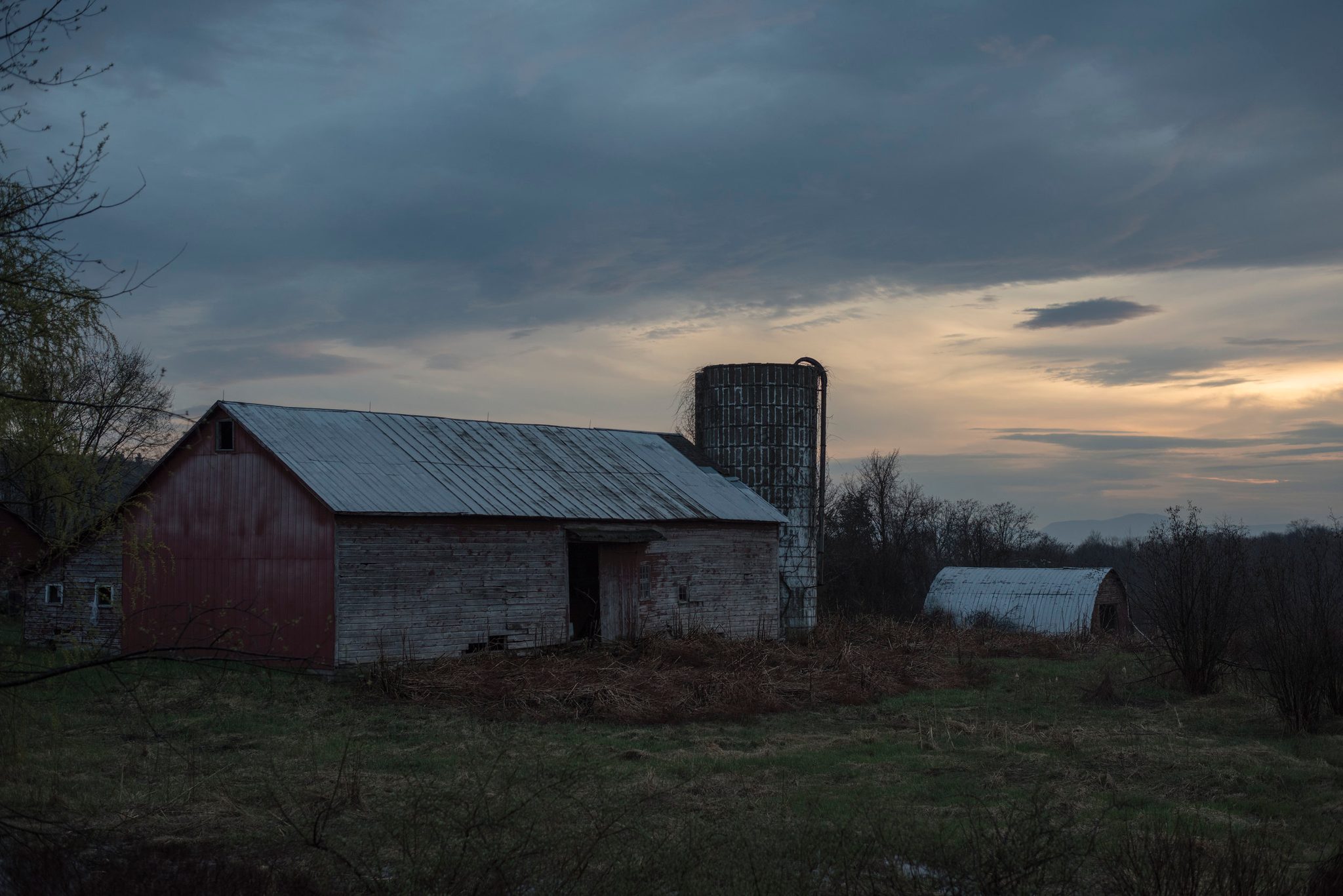 A red barn and silo stand under a heavy sky as the sunset breaks through.