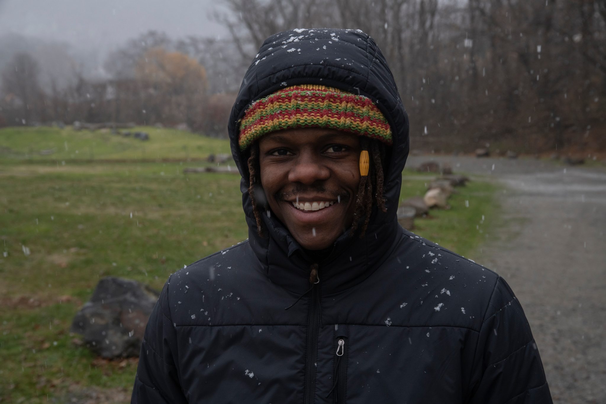 Hood up and snow on his jacket, Chev breaks into a grin in a winter field.