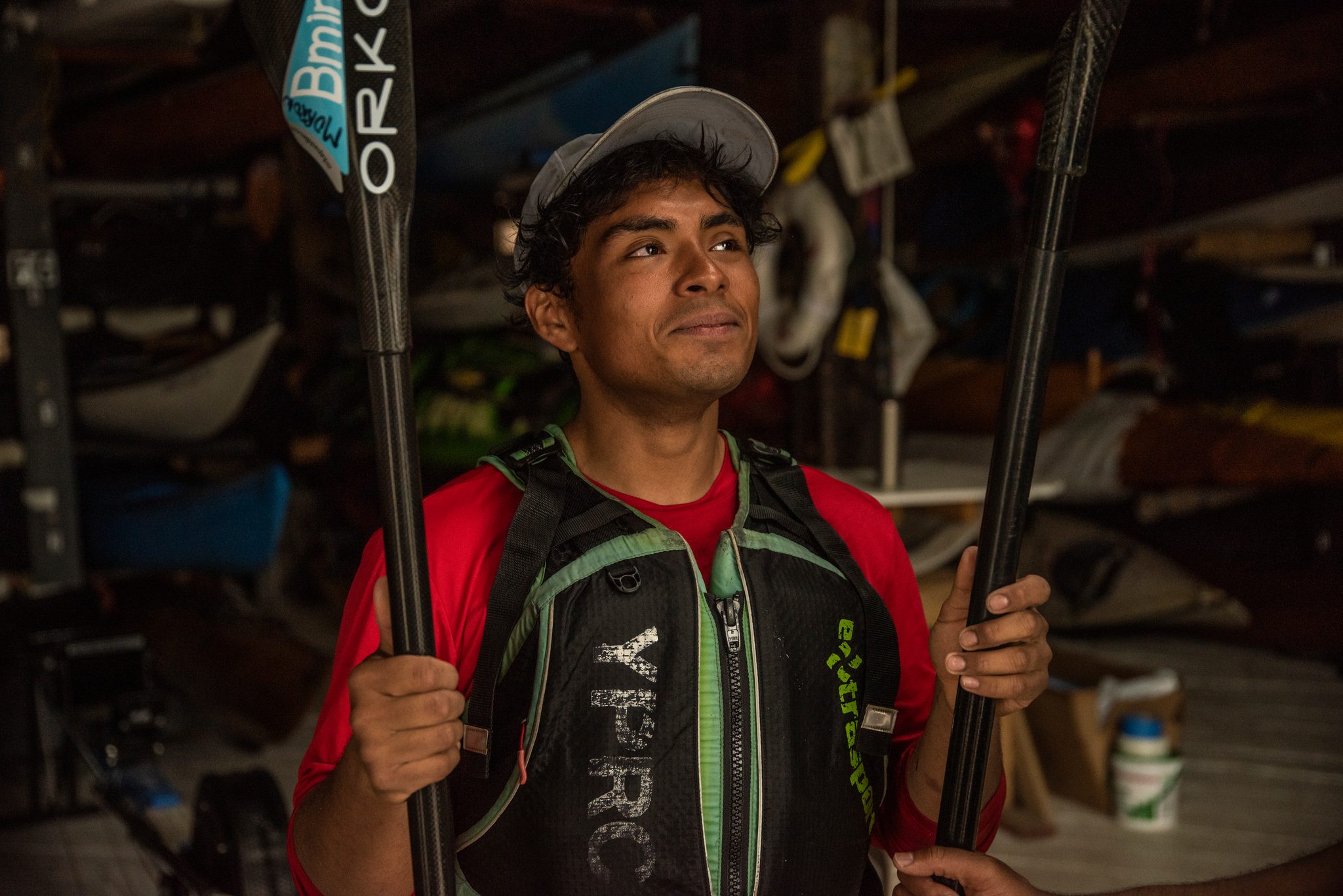 A young paddler holds his paddle upright among the racked boats, eyes on something off to the side.