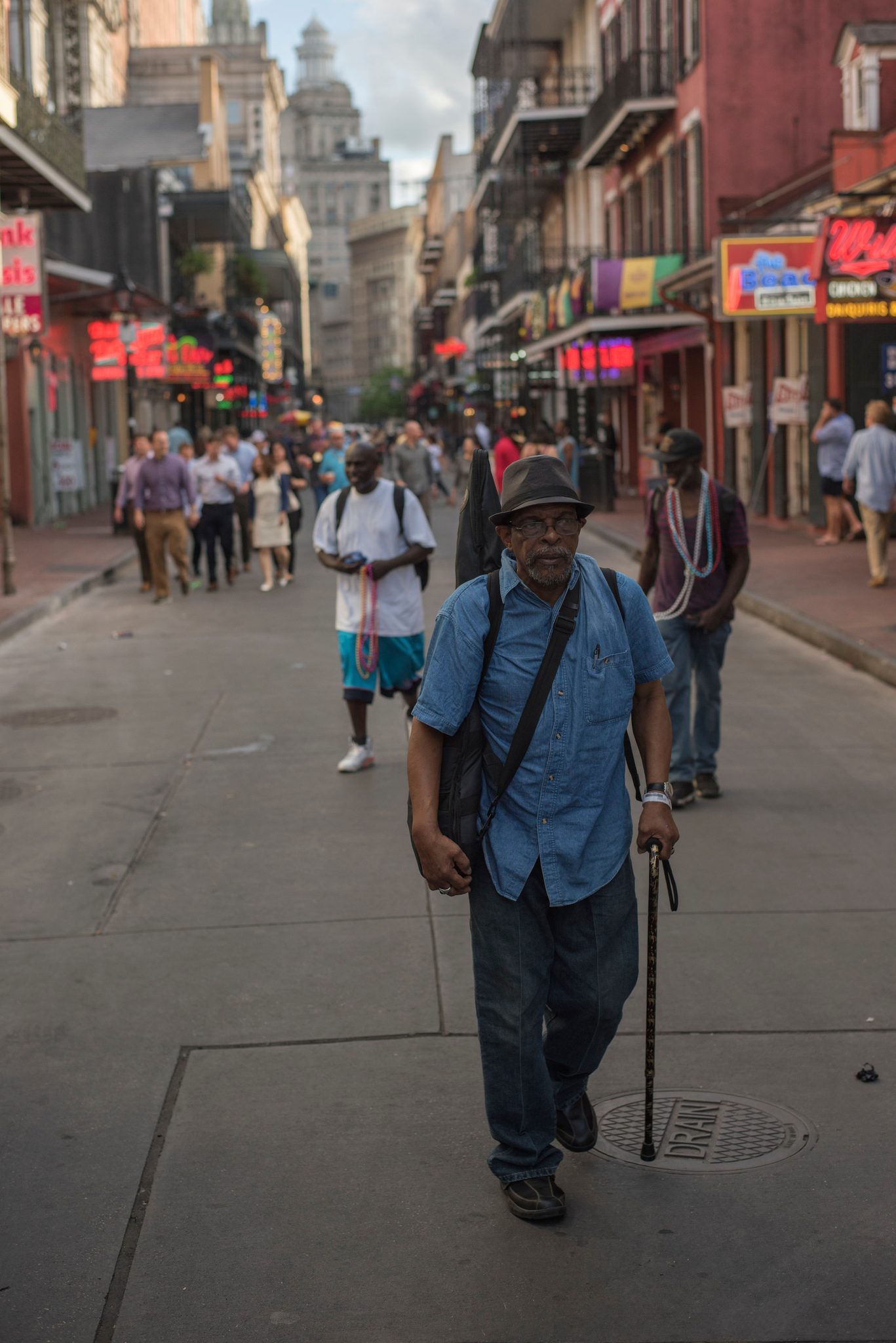 A man with a cane walks Bourbon Street as the neon comes on. There are old souls that walk these roads.