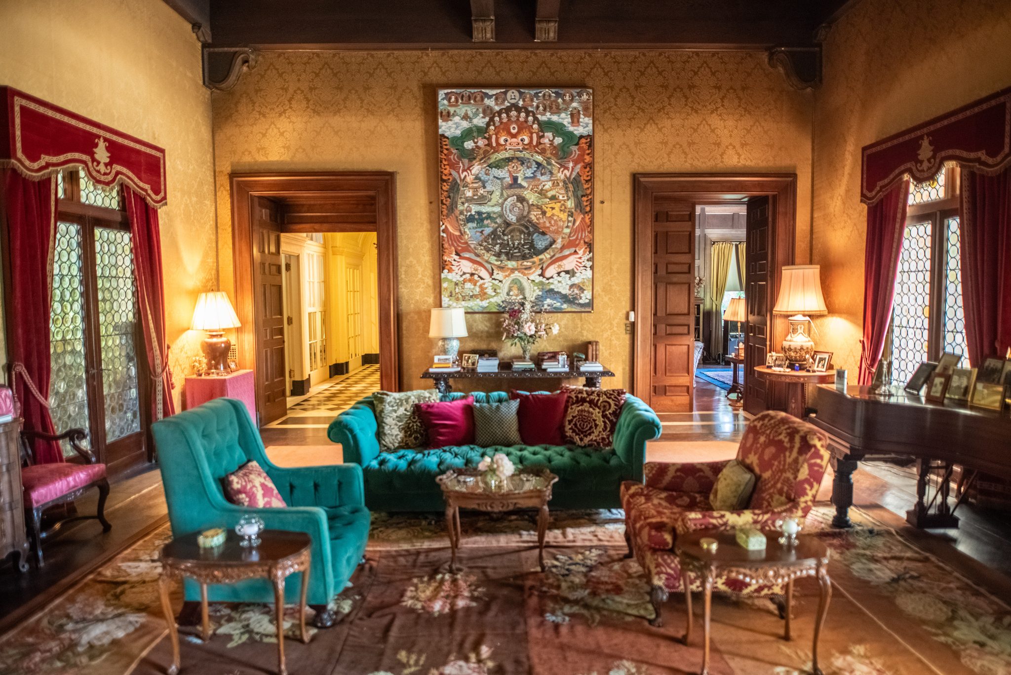 A sitting room of gold damask walls and red velvet curtains, a teal chesterfield below a large painted scroll.