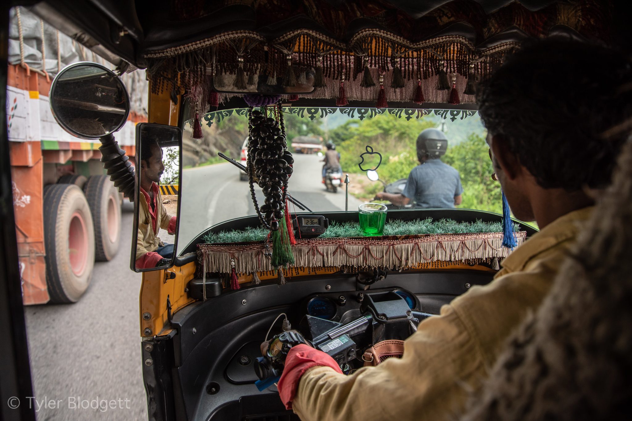 From the back seat of an auto rickshaw, the road climbs into green hills with a truck close enough to touch.