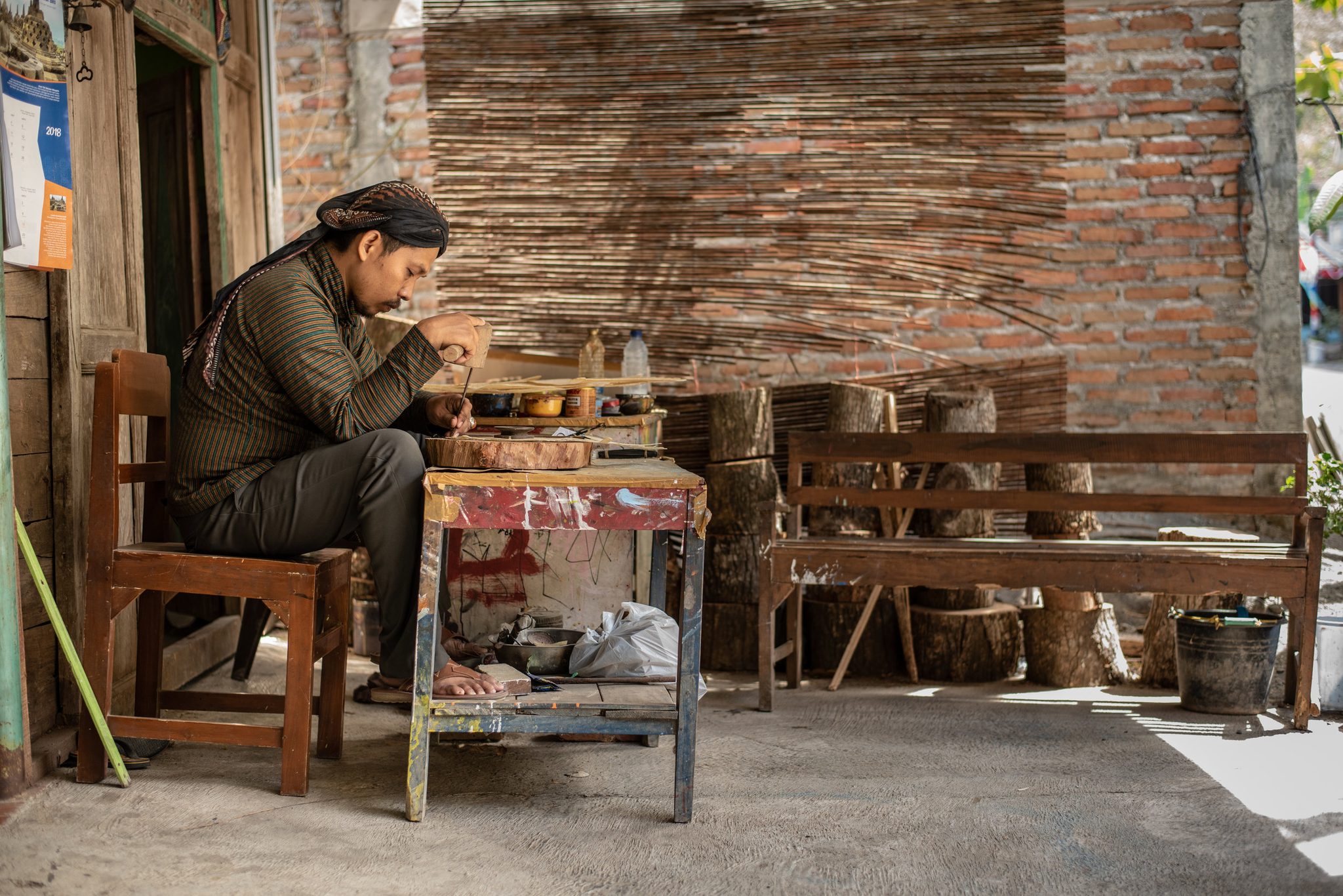 He works alone at a small table in the courtyard, the light falling through the woven wall.