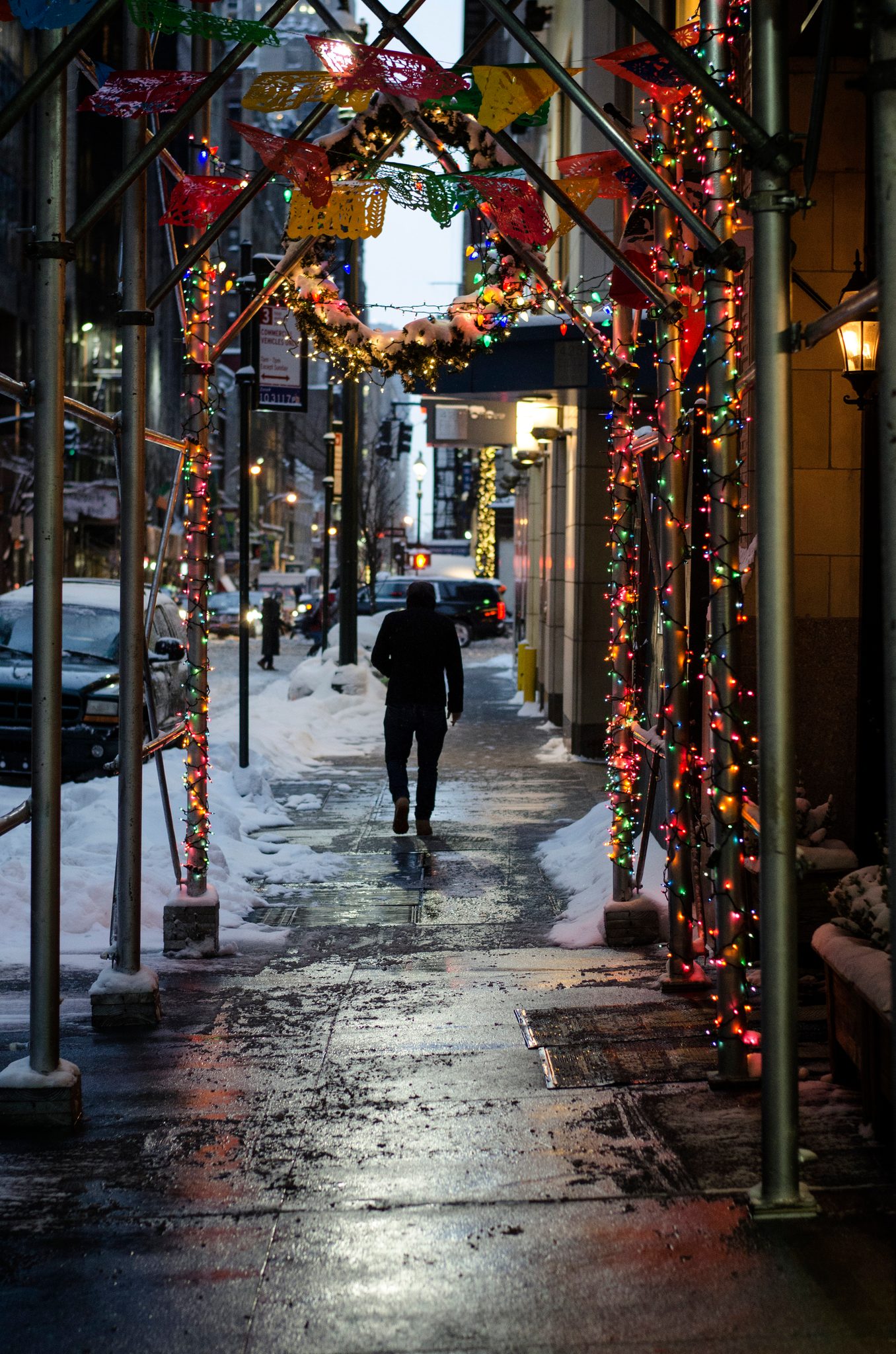 A lone walker passes beneath scaffolding strung with holiday lights, the snow gone to slush.