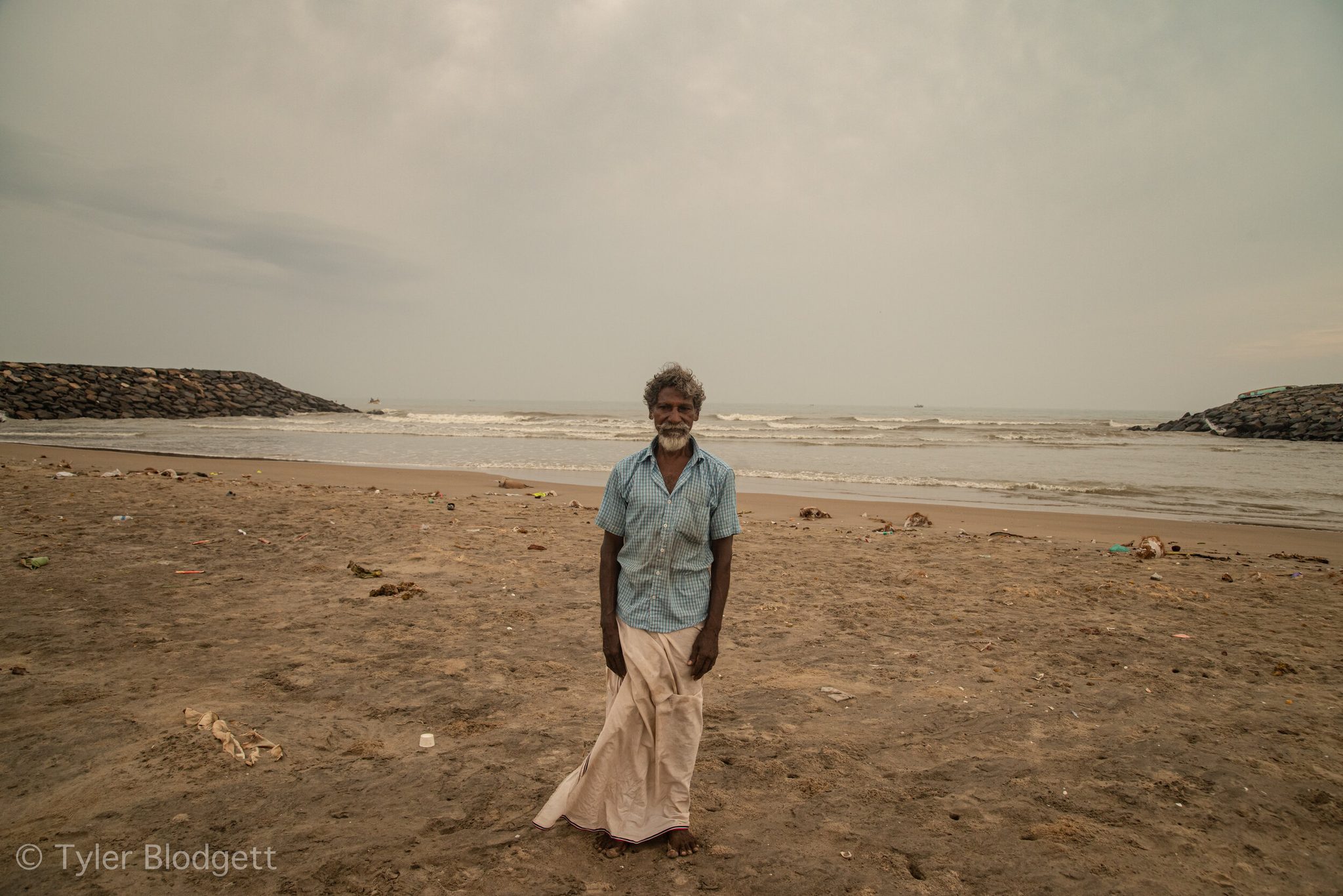 An elder in a white lungi stands on the beach between the breakwaters, the sea gray and restless behind him.