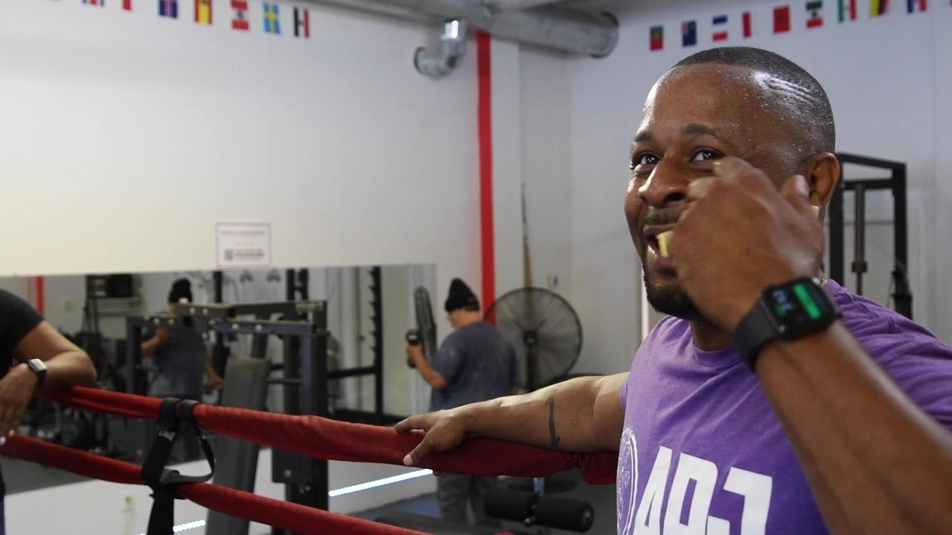 At the ropes in a club shirt, a trainer throws a mock punch at the camera and laughs.