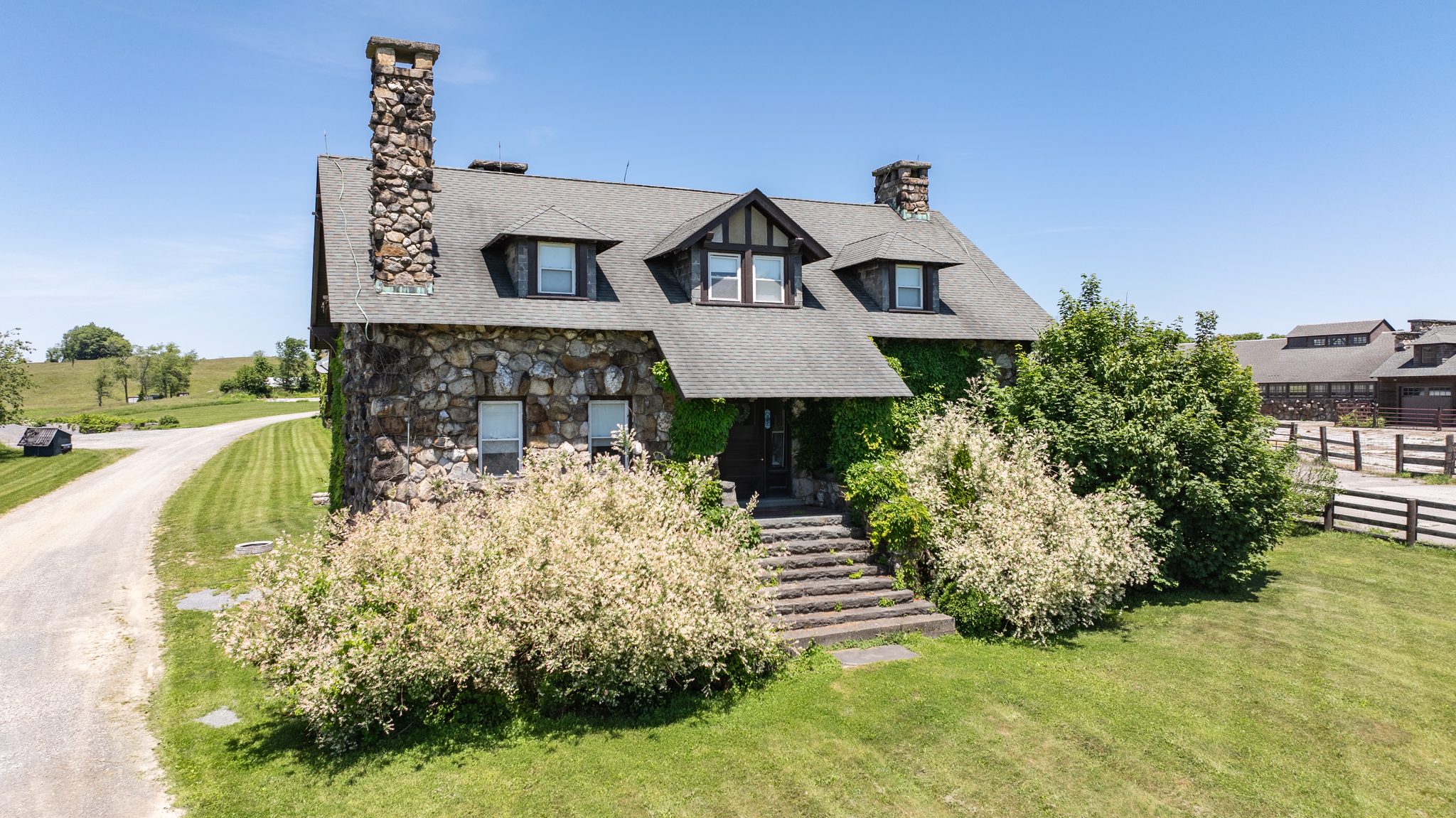 A stone cottage by the barns with a fieldstone chimney, three dormers and flowering shrubs along the front steps.