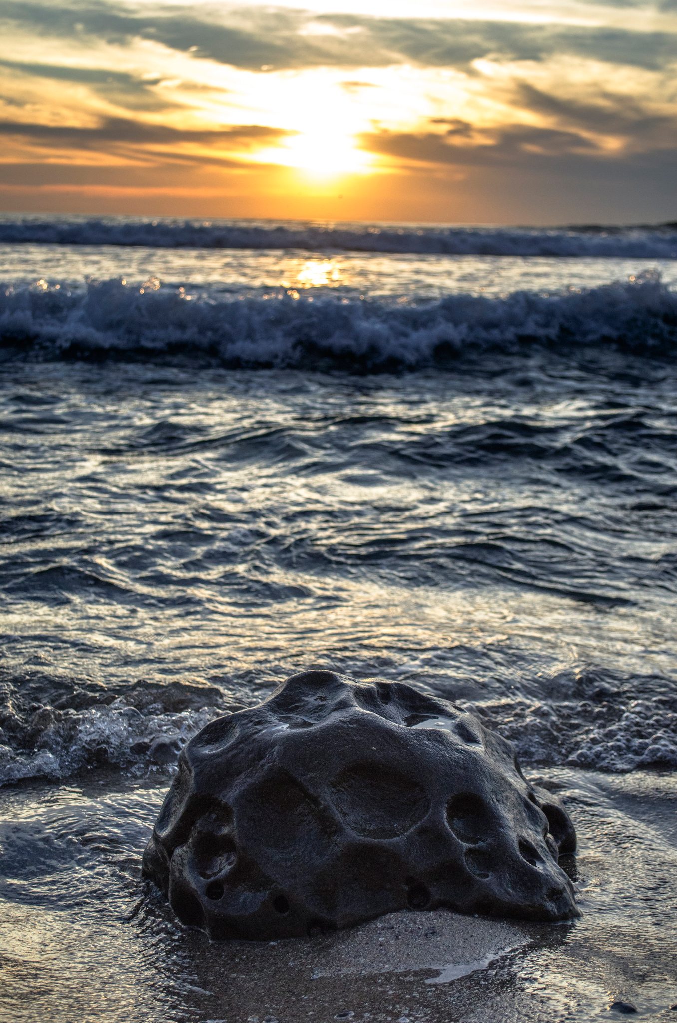 A dark stone sits in the surf as the sun sets over the waves.