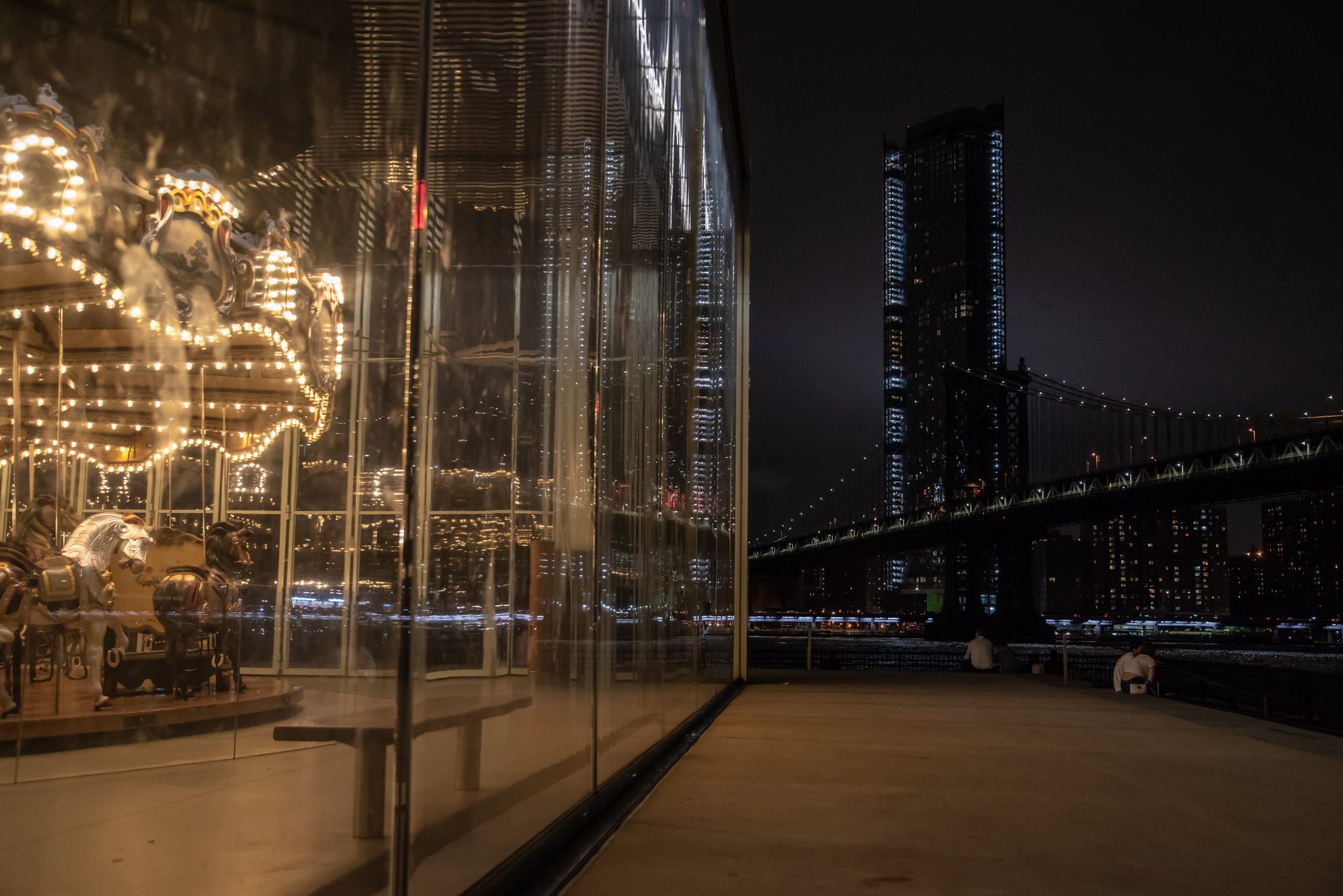 A lit carousel turns inside its glass pavilion on the waterfront, the bridge and dark towers beyond.