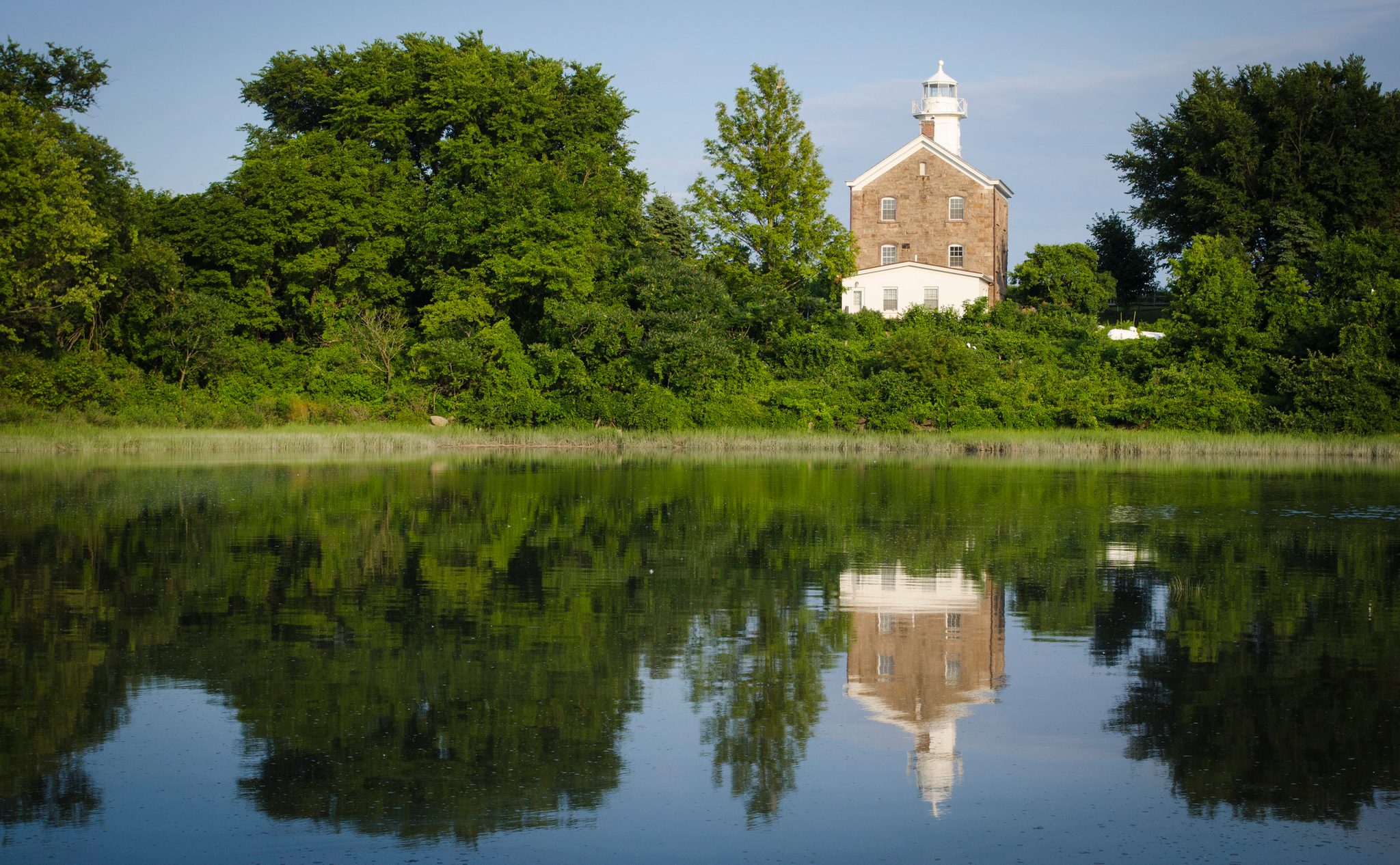A stone lighthouse rises above the trees, doubled in the still pond.