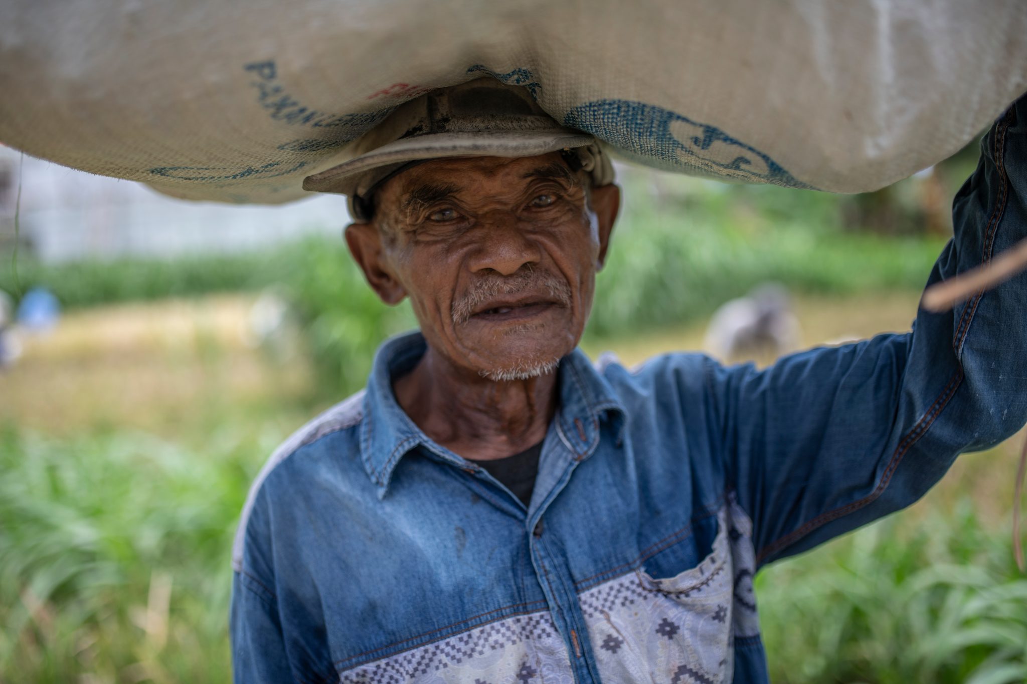 An old man carries a heavy sack on his head, his face creased by the sun.
