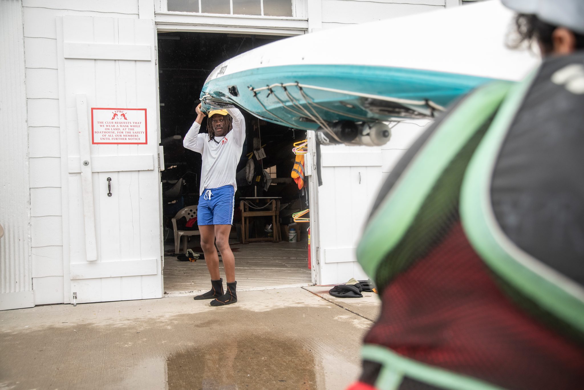 Chev carries a kayak overhead out through the boathouse doors while a paddler waits in the foreground.