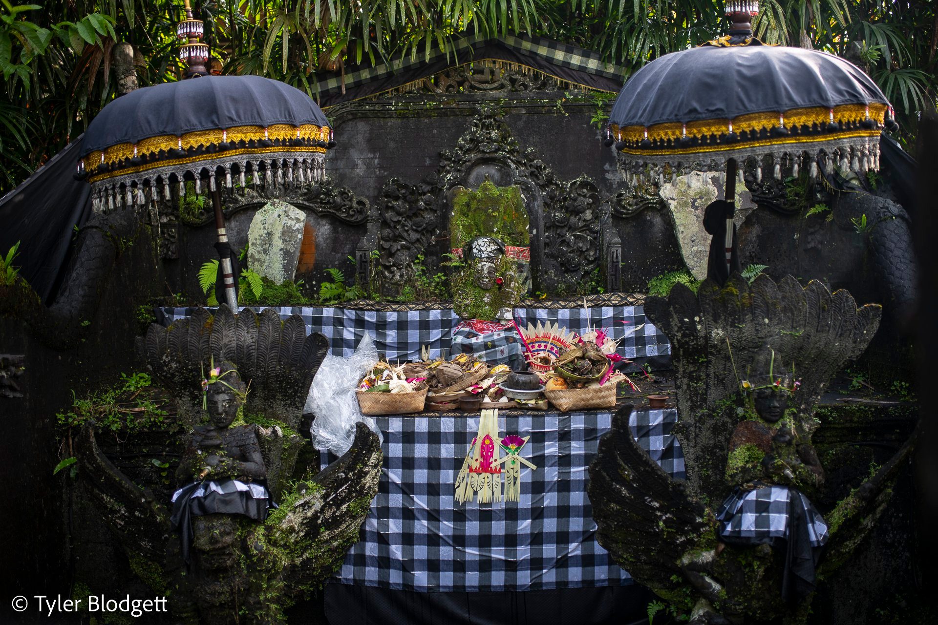 Offerings sit on the shrine beneath fringed umbrellas, the stone wrapped in black and white checked cloth.