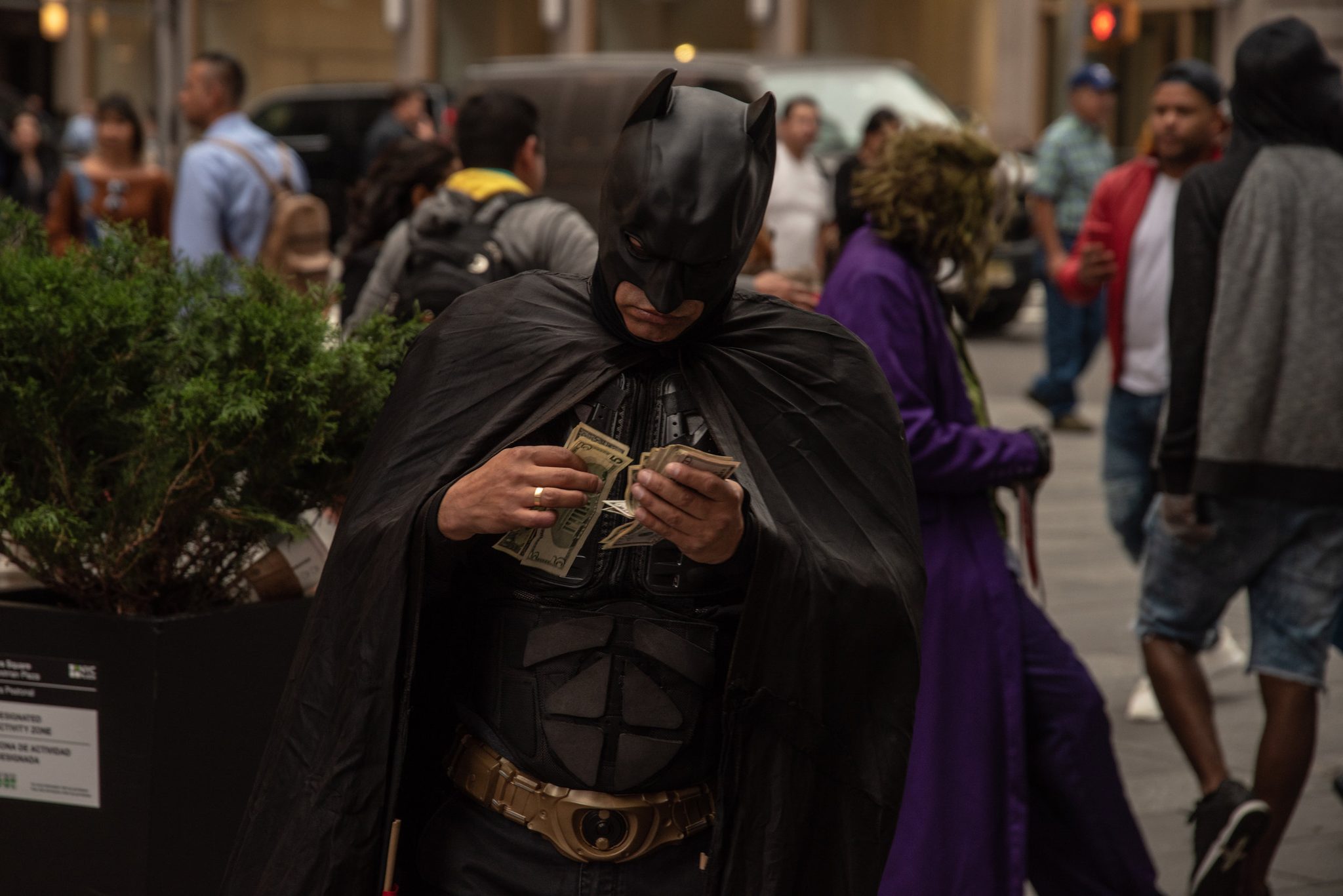 Batman counts his tips on the plaza, the Joker lurking just behind.