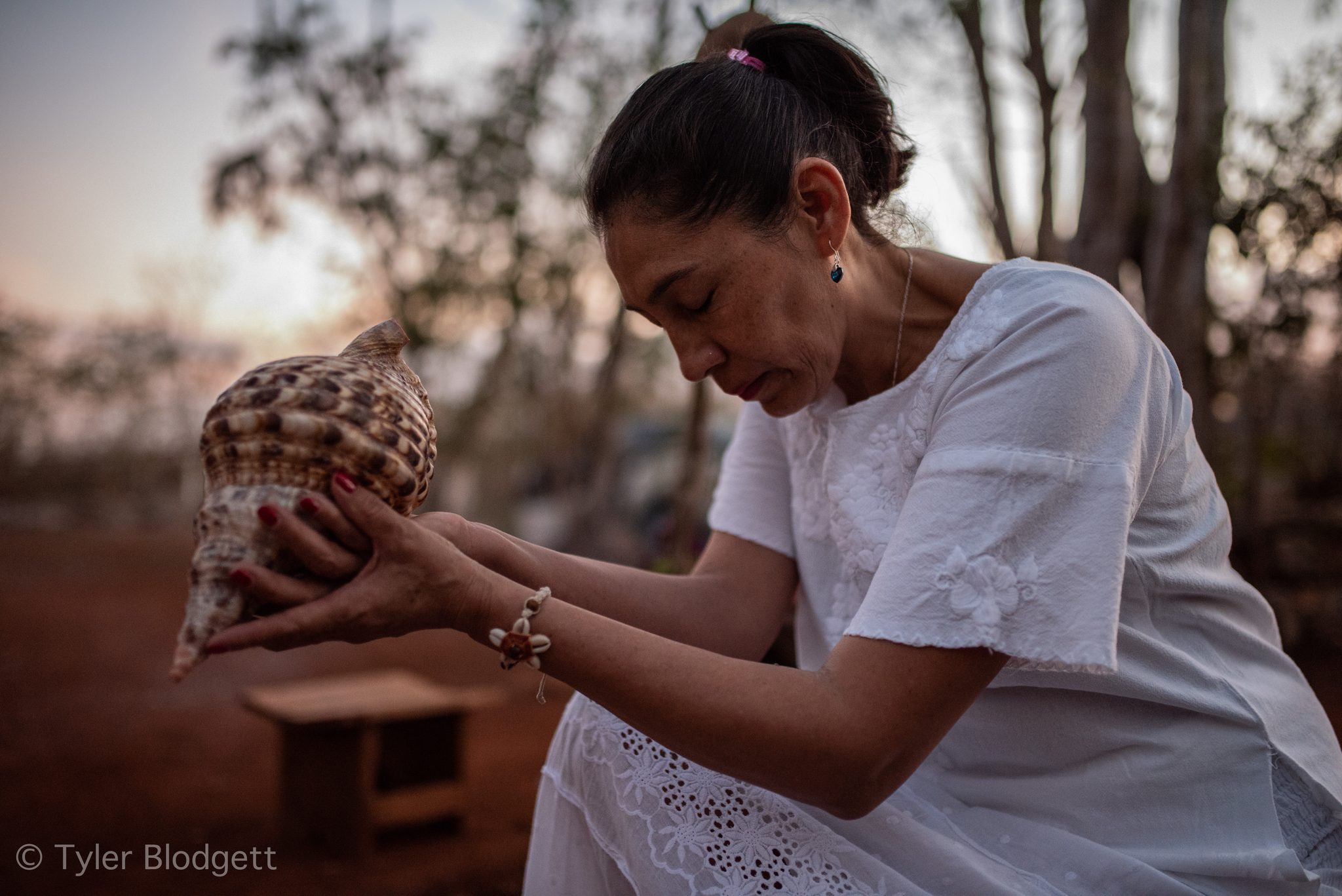 A woman in white cradles a spiral shell in both hands, eyes lowered.