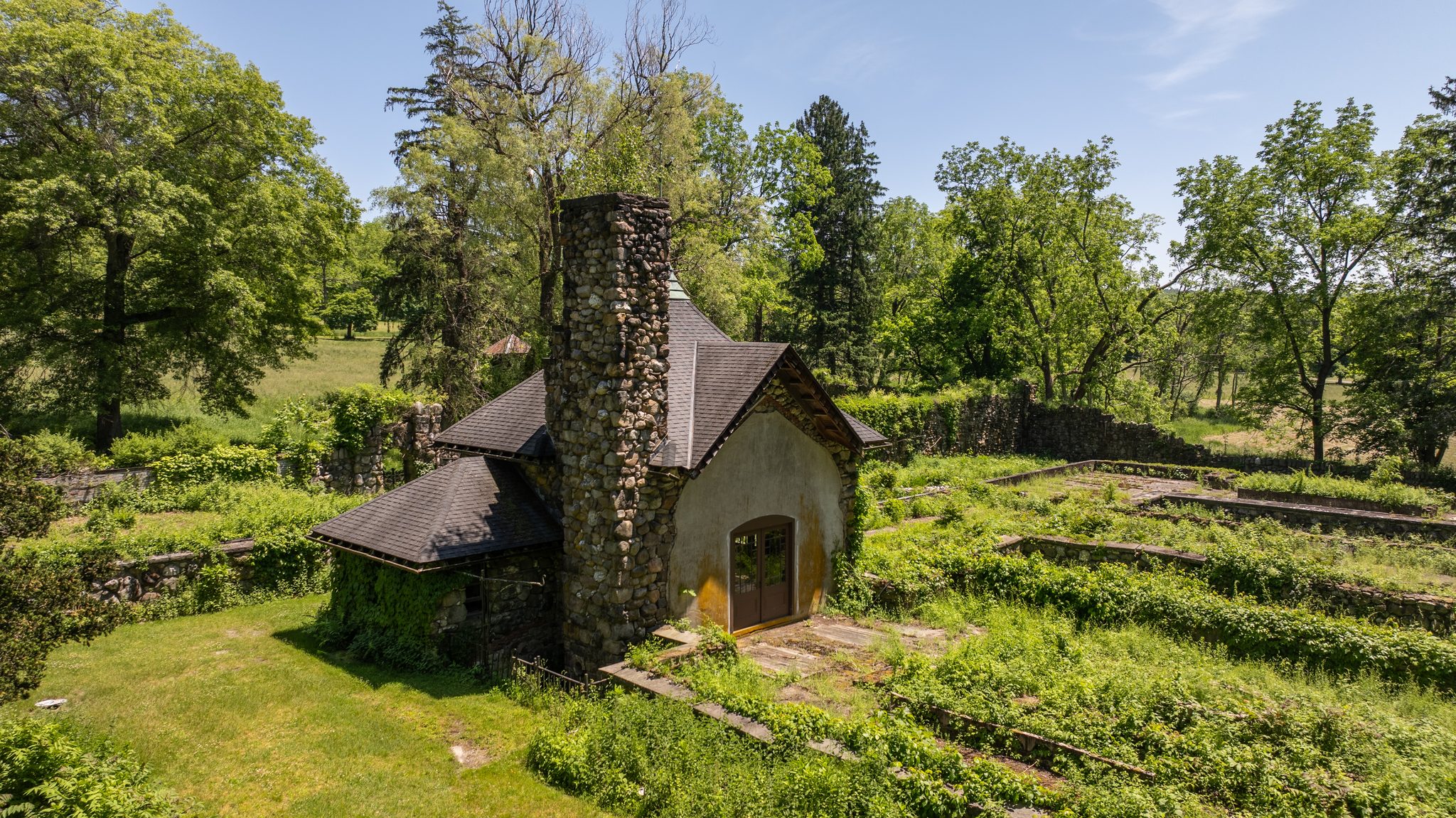 A small stuccoed cottage with a fieldstone chimney stands among overgrown garden walls and tall trees.