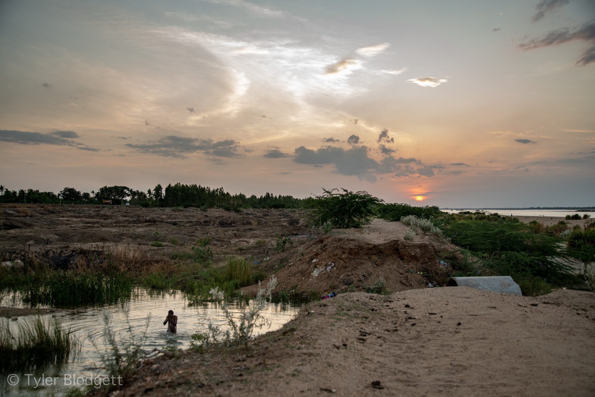 The sun sets over a riverbed of sand and shallow pools, a lone figure wading.