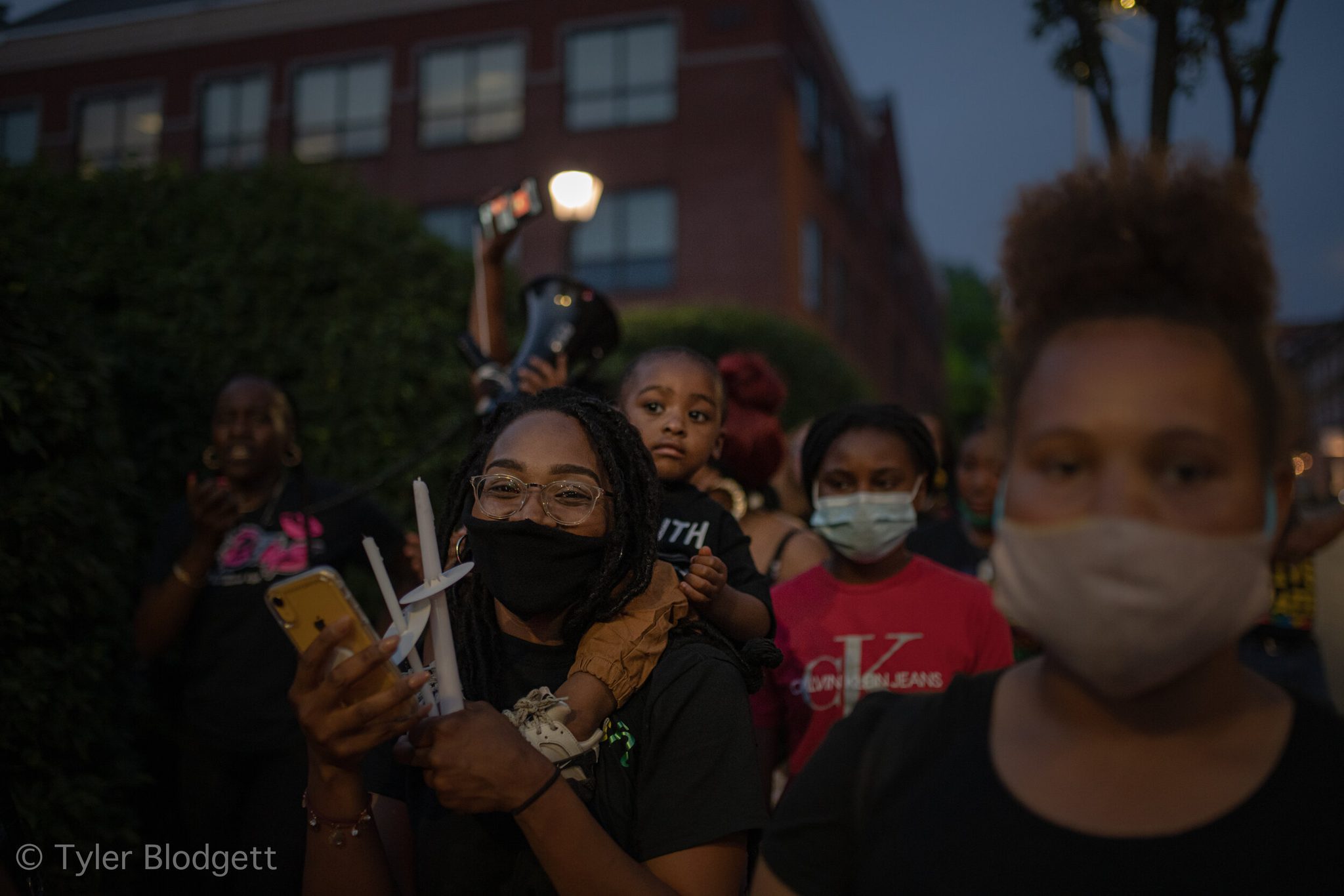 Women and children gather with candles, a little one riding high on someone's shoulders.